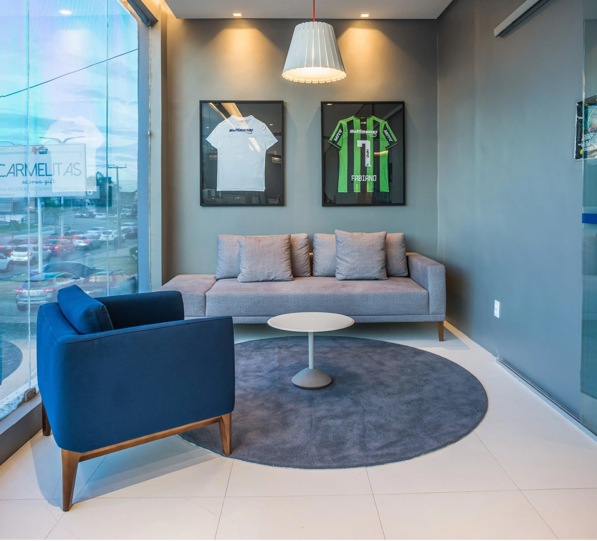 Modern interior with a beige sofa, a dark blue armchair, a round gray rug, a small white coffee table, framed soccer jerseys on the wall, and large windows showing a parking lot outside.