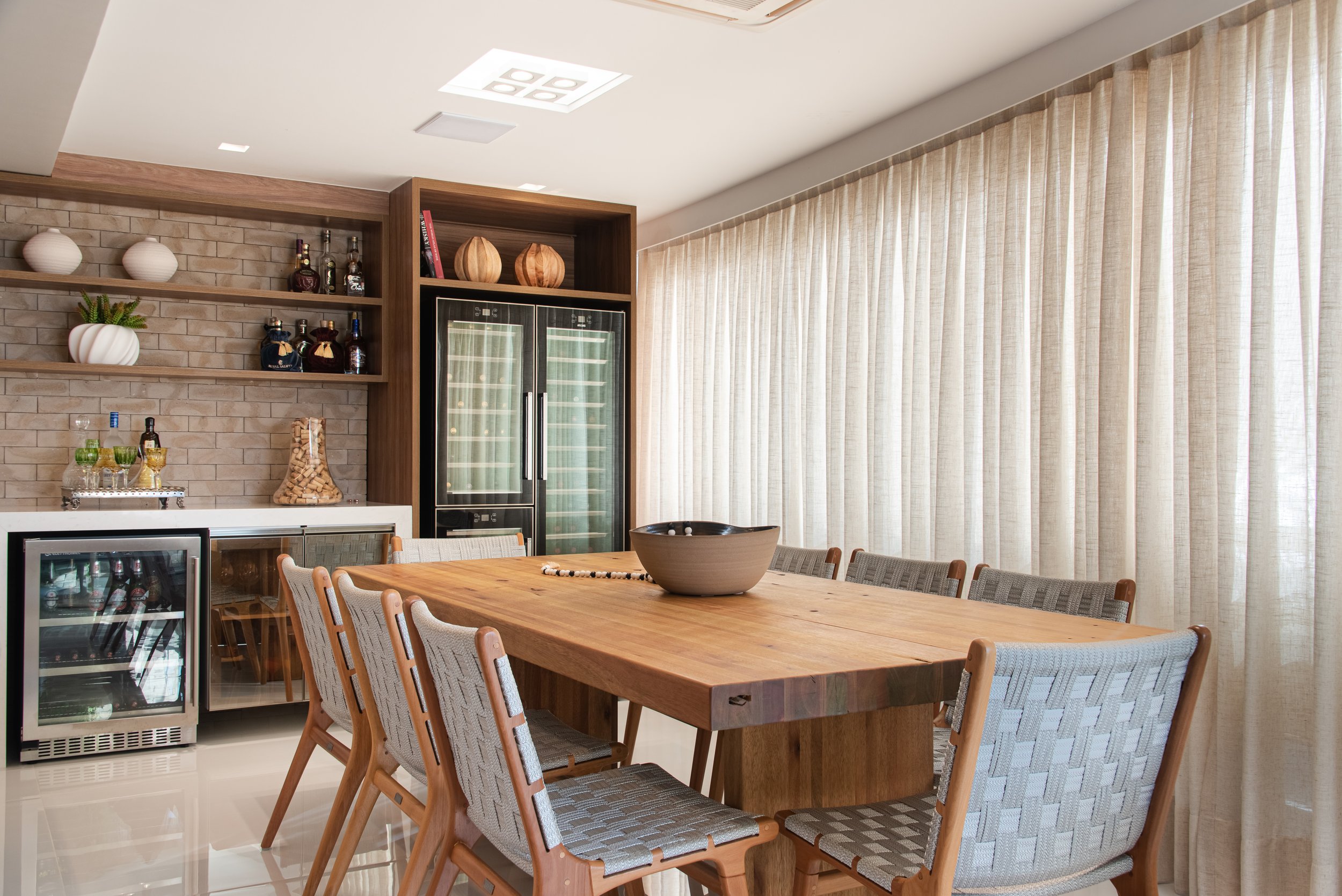 Modern dining room with a large wooden table surrounded by upholstered chairs, a large bowl on the table, and a backdrop of large beige curtains covering windows. There is a built-in espresso bar with bottles, a wine fridge, and decorative shelves.
