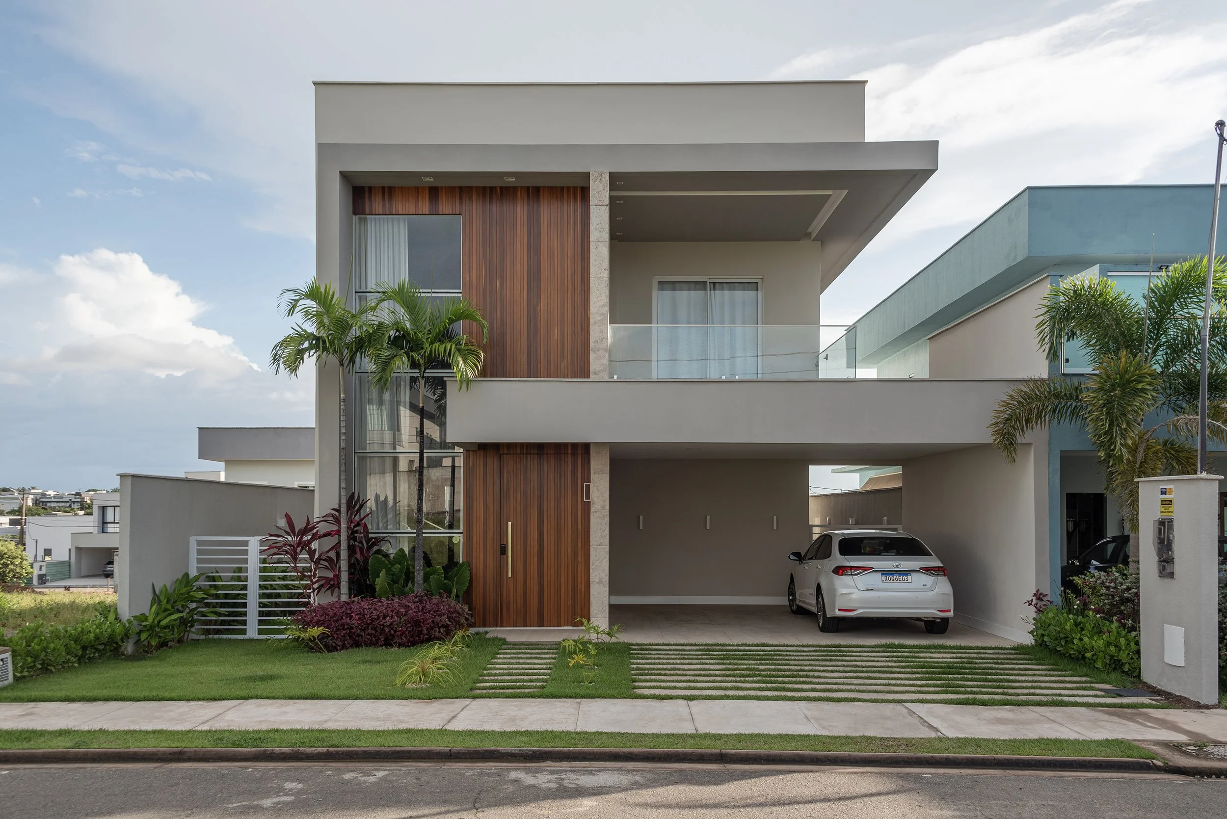 Modern two-story house with a car parked in the garage, wooden and concrete exterior, tropical plants, and a grassy front yard.
