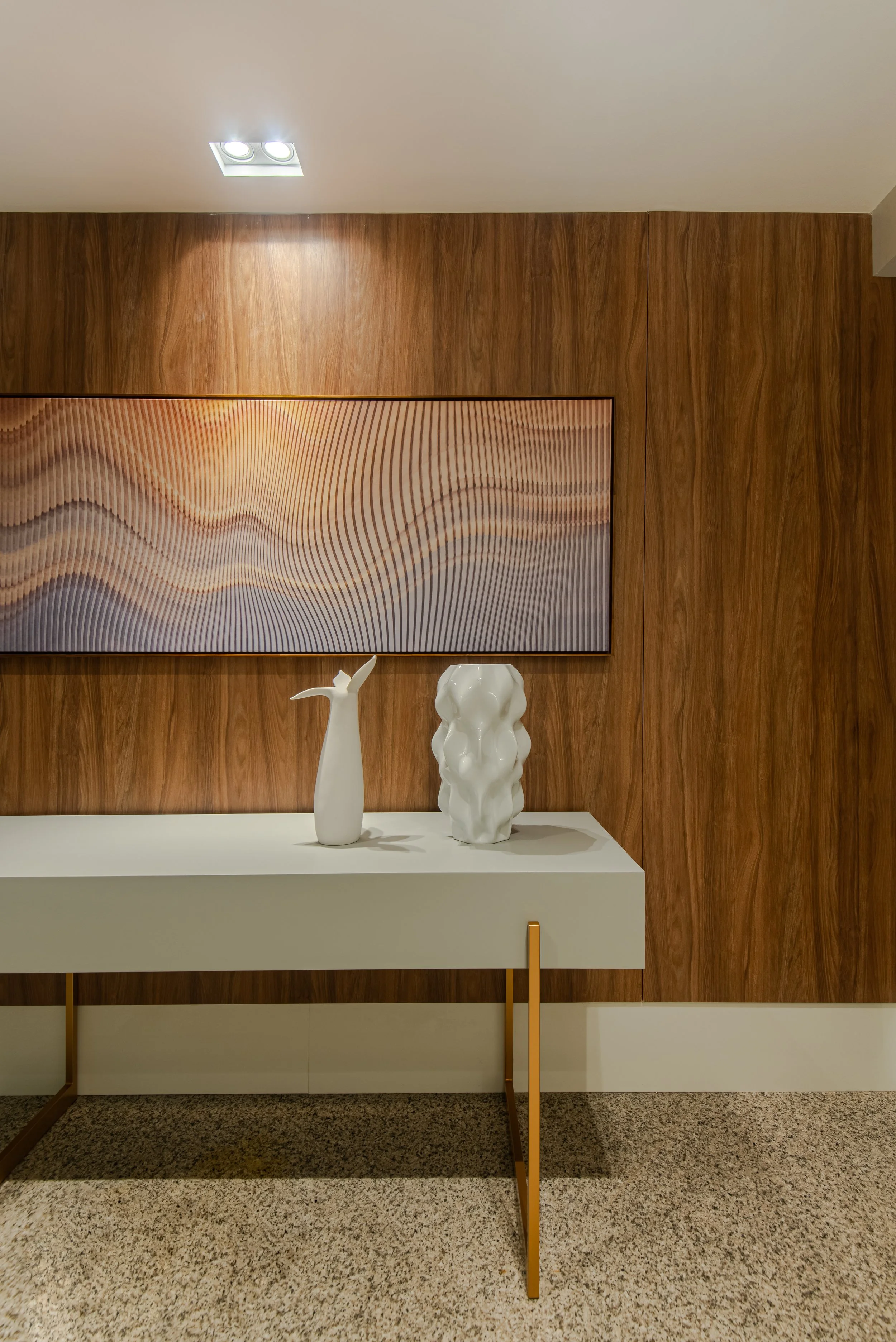Interior wall with a wood panel finish, two white sculptural vases on a white console table, a rectangular abstract art piece or photograph on the wall, ceiling lights, carpeted floor.