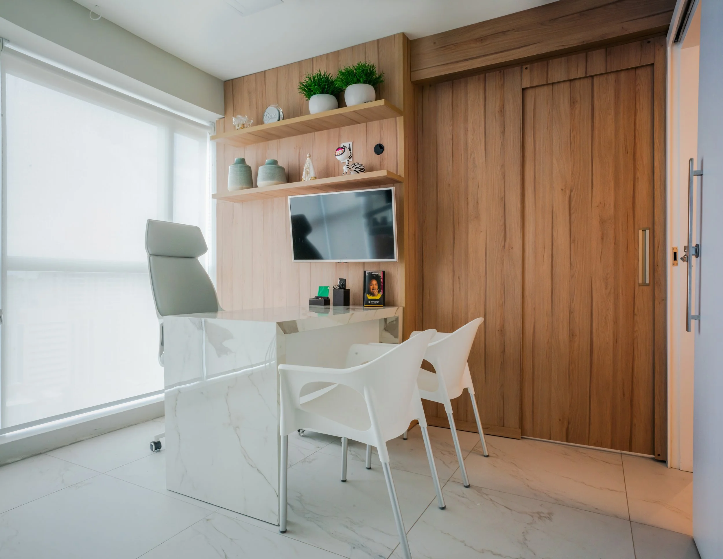 Modern office workspace with white marble desk, gray high-backed chair, two white plastic chairs, wooden wall with shelves, potted plants, decorative items, wall-mounted TV, and large window with white blinds.