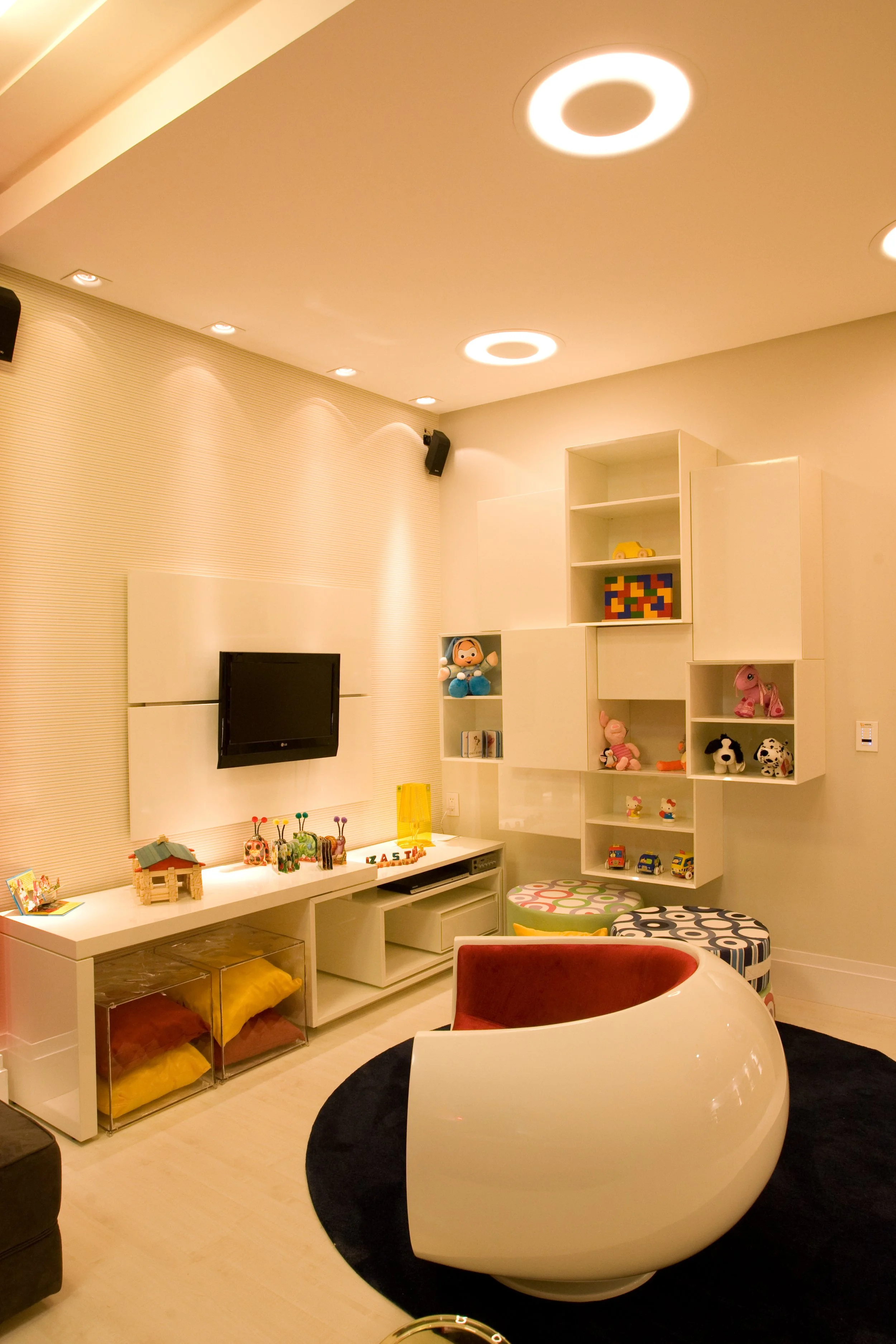 A modern living room with a white entertainment unit, a wall-mounted flat-screen TV, and a cozy white and red swivel chair. The room features built-in shelves with toys and decorative items, ceiling circular lights, and a black circular rug on light-