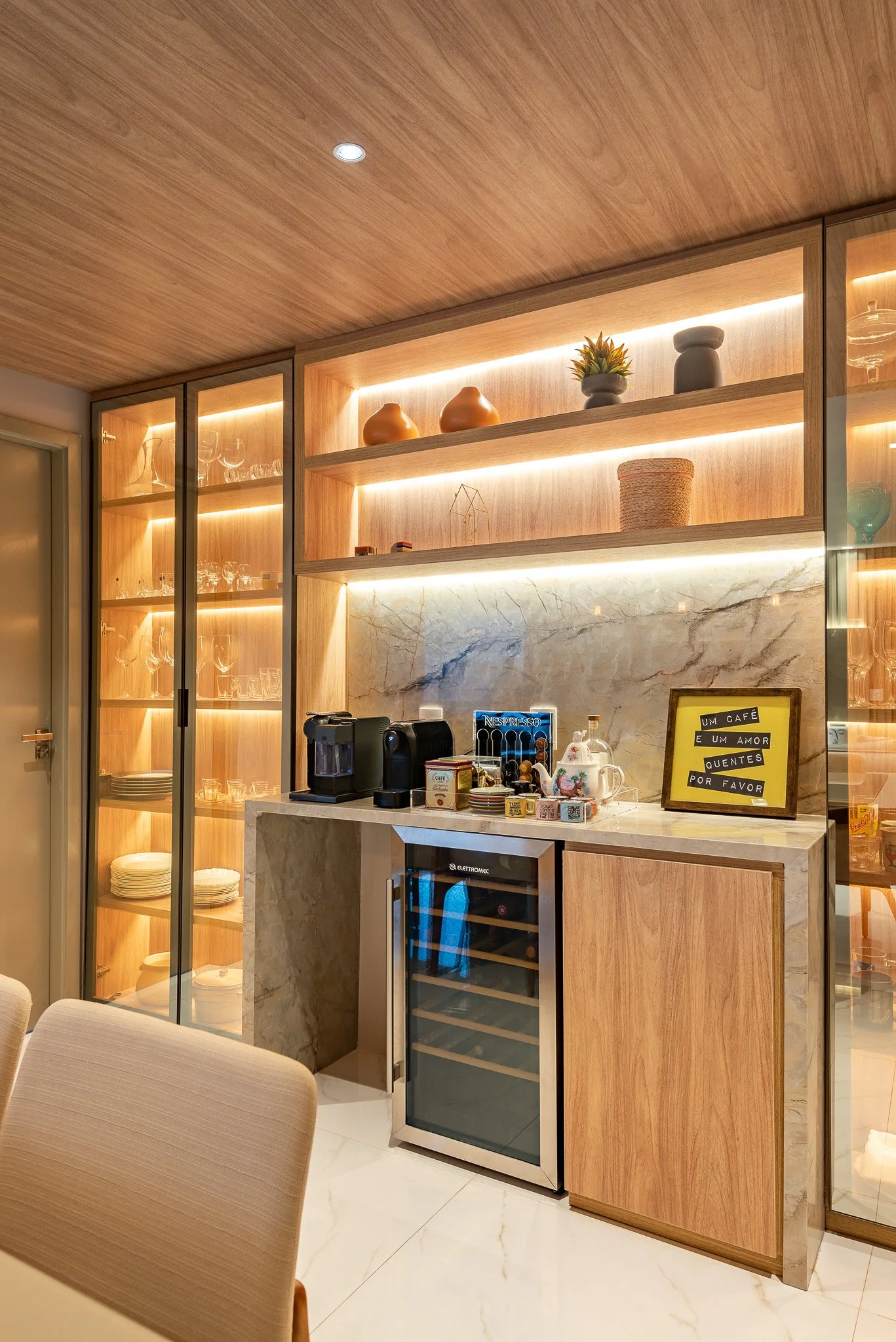 Coffee station with a small wine refrigerator, coffee machine, cups, and a framed sign in front of a marble backsplash, surrounded by wooden shelving and glass cabinets displaying glassware and dishes.