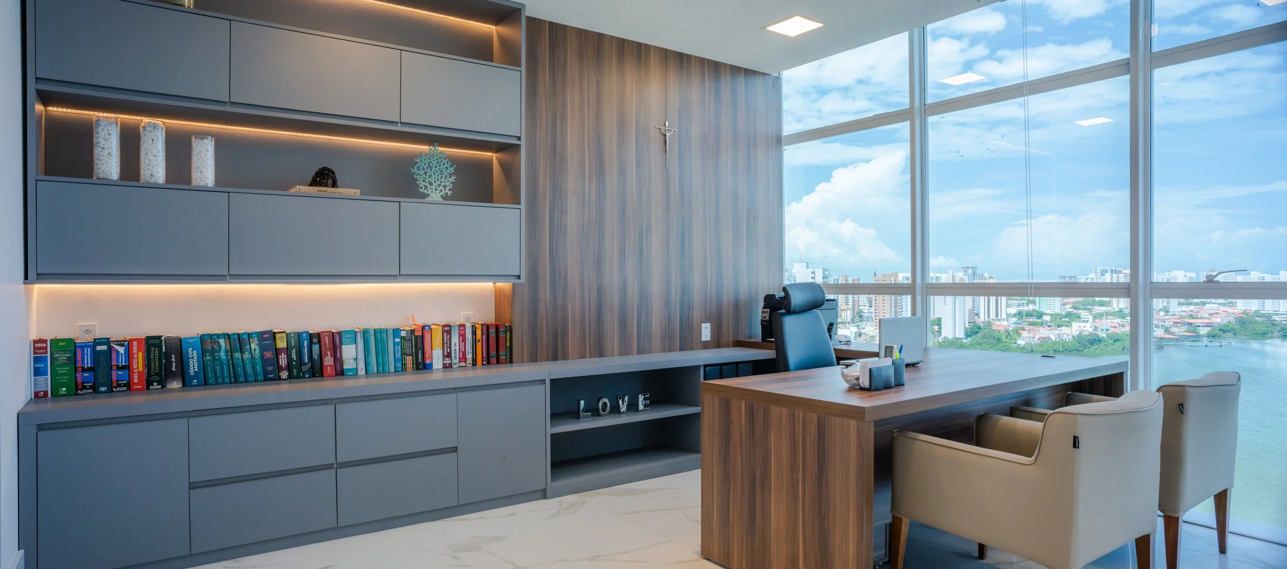 Modern office with large windows showing city skyline, wooden desk, cream chairs, bookshelf with books and decorative items, wall with wood paneling, and cross on wall.