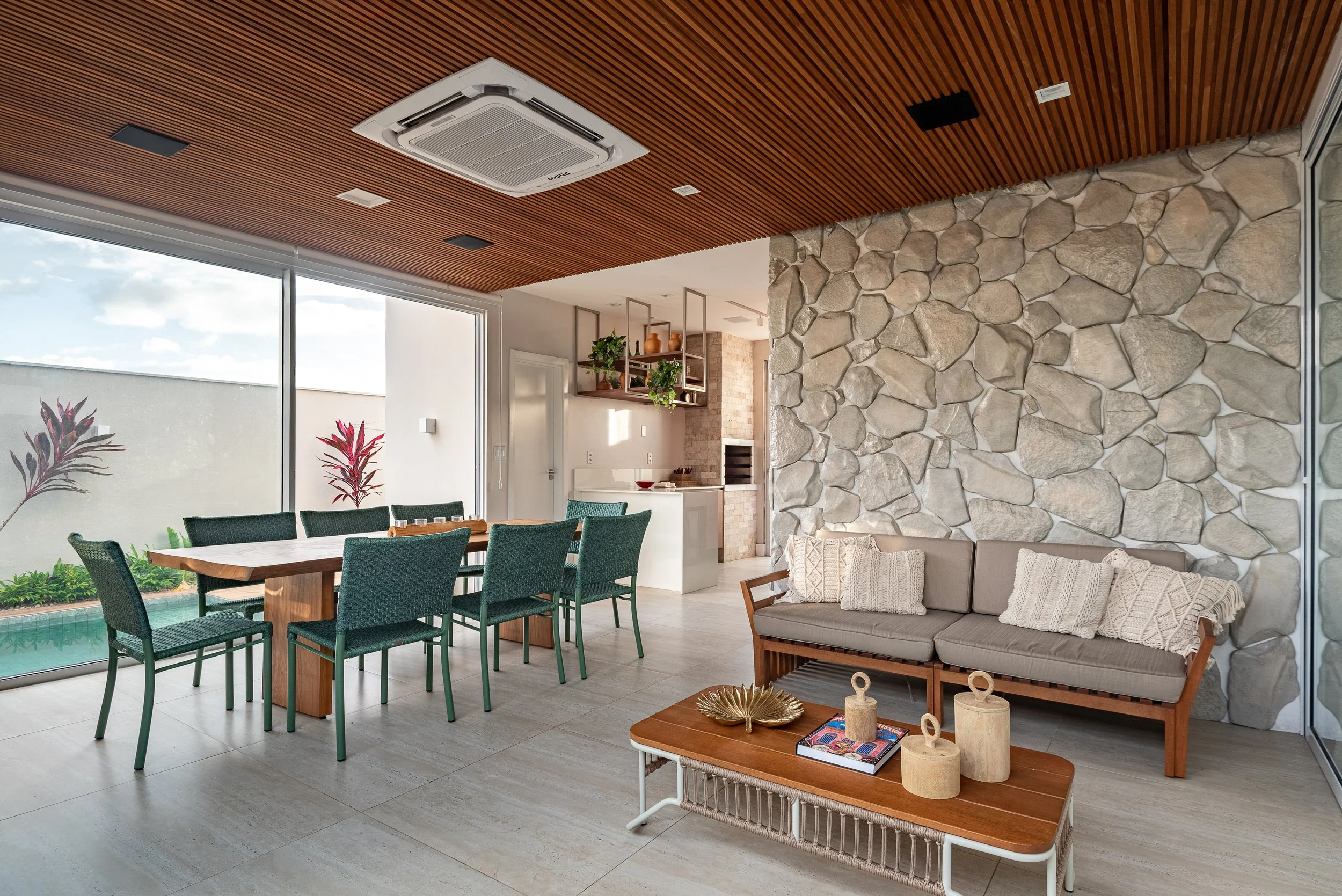 Modern indoor living space with a stone accent wall, a beige sofa with pillows, a wooden coffee table with decorative items, a dining table with green chairs, and large windows with outdoor views.