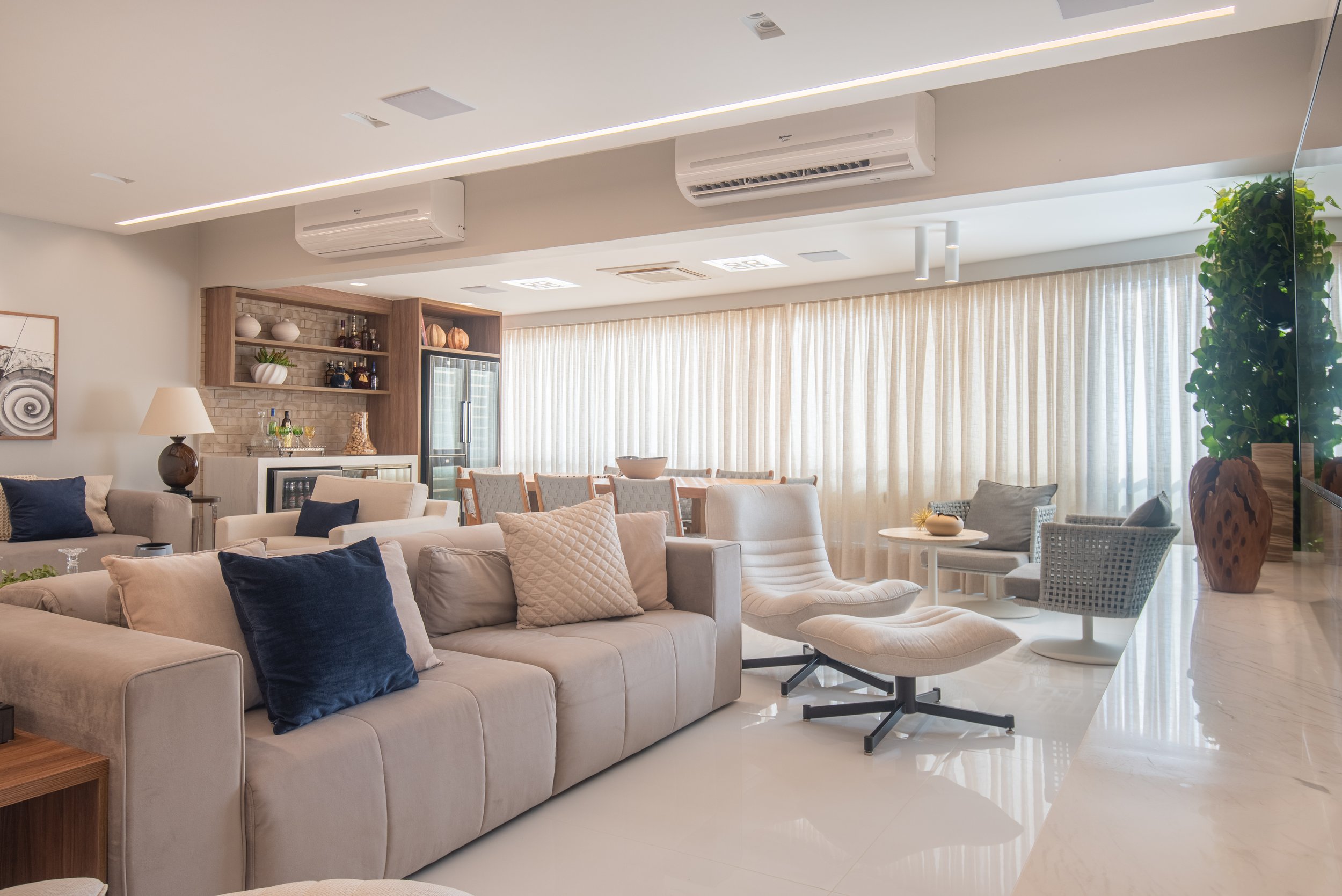 Spacious and modern living room with beige couches, armchairs, decorative pillows, a dining table with chairs, and large windows with sheer curtains, featuring minimalist decor and a bright ambiance.