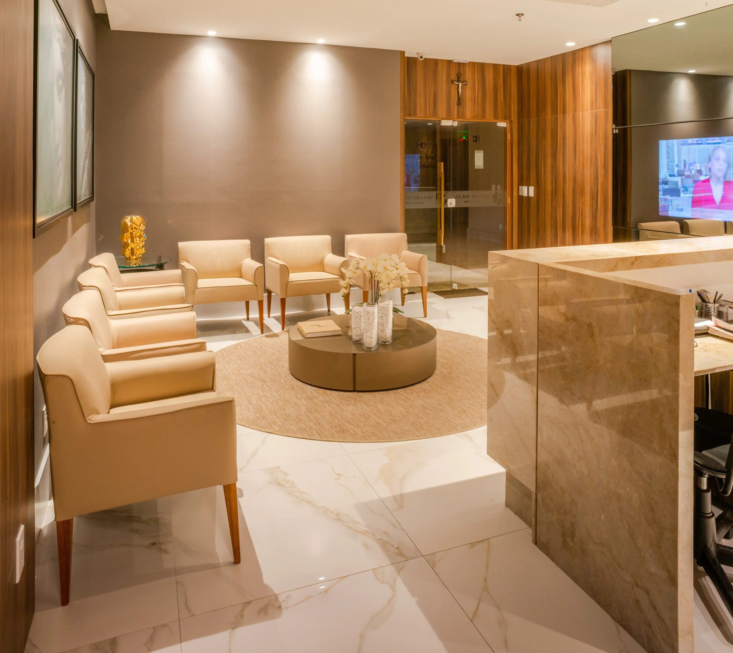 A hotel lobby with beige chairs arranged around a round table with white flowers, a small book, and decorative vases. There is a beige rug in the center, marble flooring, a wooden wall with a crucifix, a glass door, and a television showing a news an
