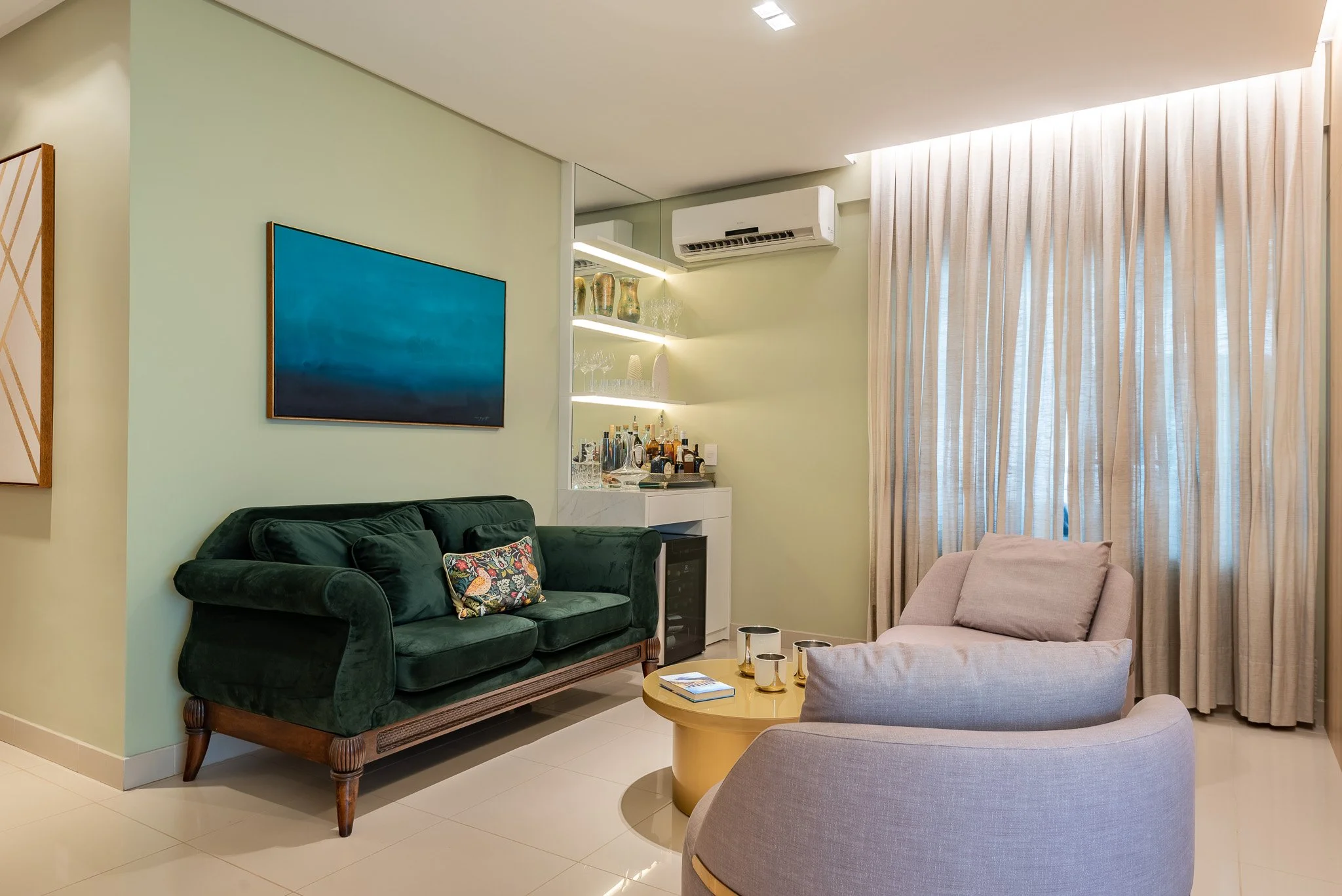 Living room with green velvet sofa, beige armchair, light green walls, beige curtains, wall-mounted TV, bar area with bottles and glasses, and a small round wooden coffee table.