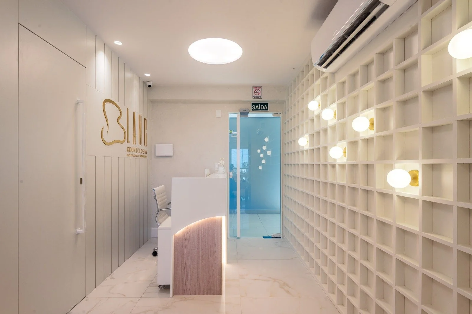 Modern dental clinic reception area with white decor, illuminated wall lights on a grid wall, and a glass door labeled 'SAÍDA' (exit).