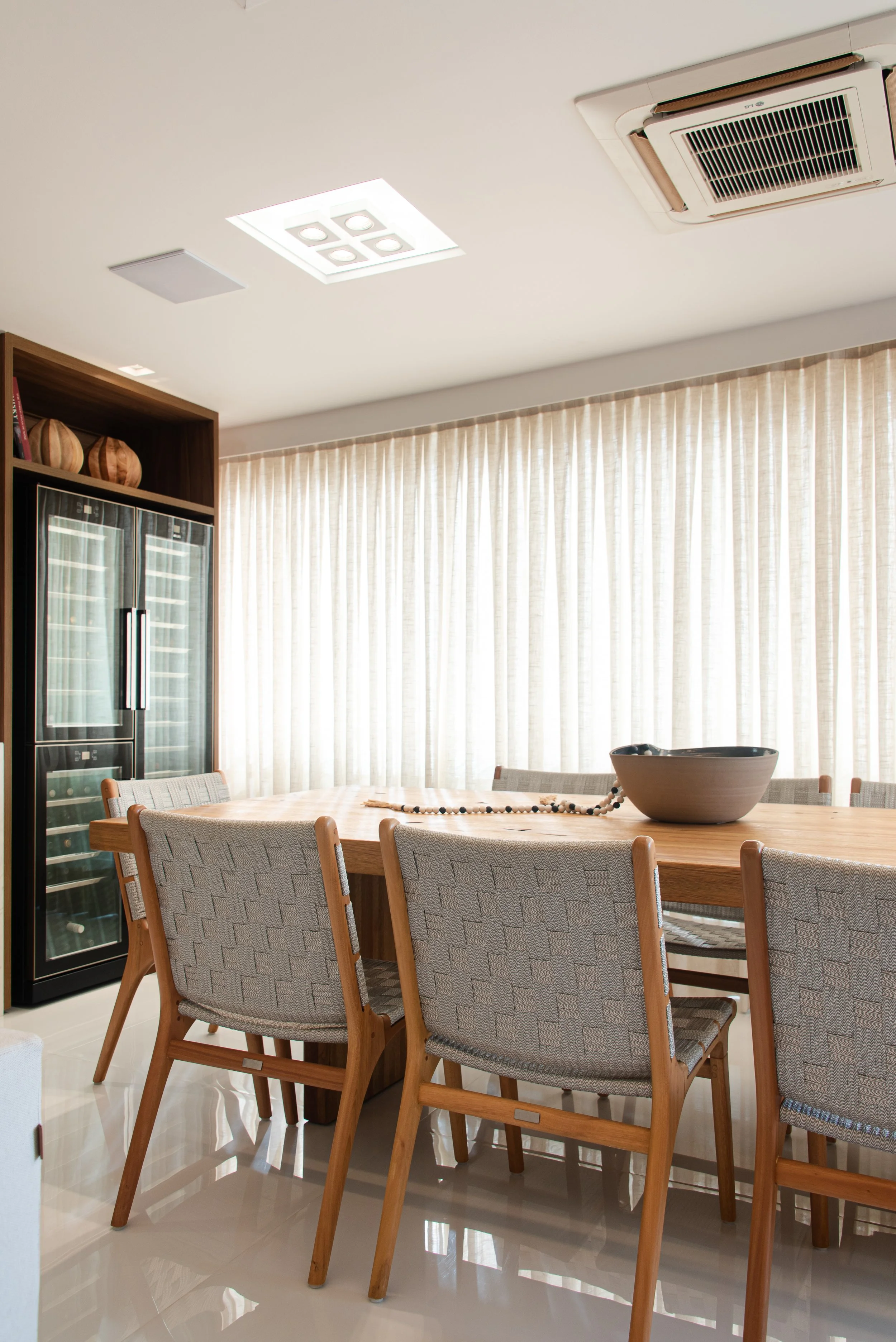 Dining room with a large wooden table, six upholstered chairs, a beige curtain, a black beverage cooler, decorative vases on a shelf, a black bowl on the table, and a ceiling with recessed lights and an air conditioning unit.