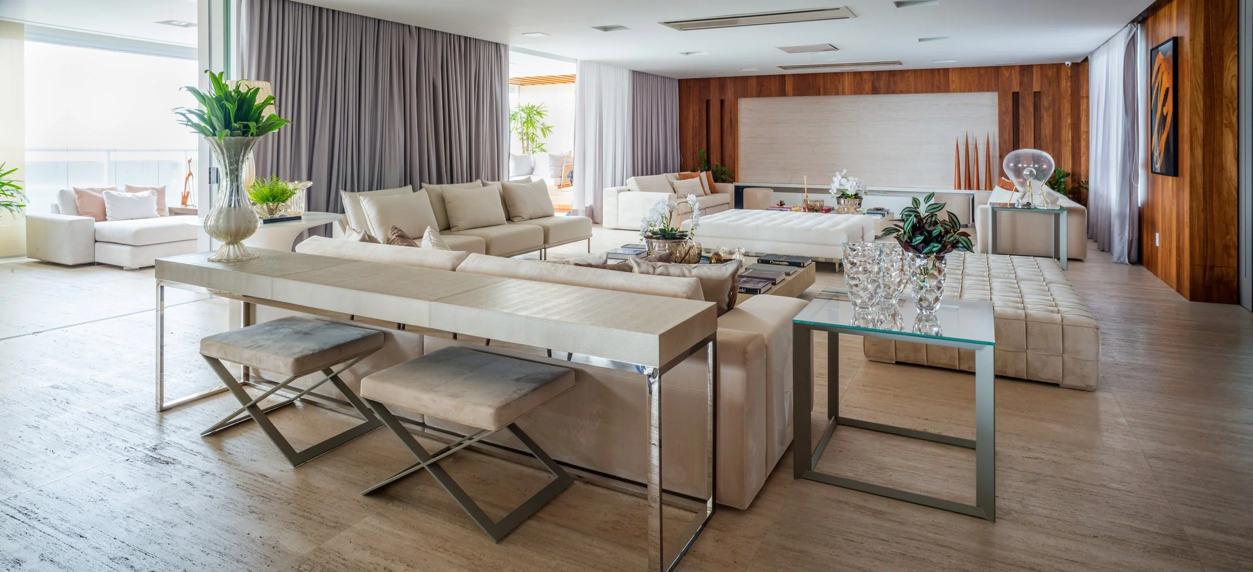 Modern living room with white sofas, glass tables, and wooden wall decor. Large windows with curtains and potted plants. Decor includes vases with flowers and abstract art.