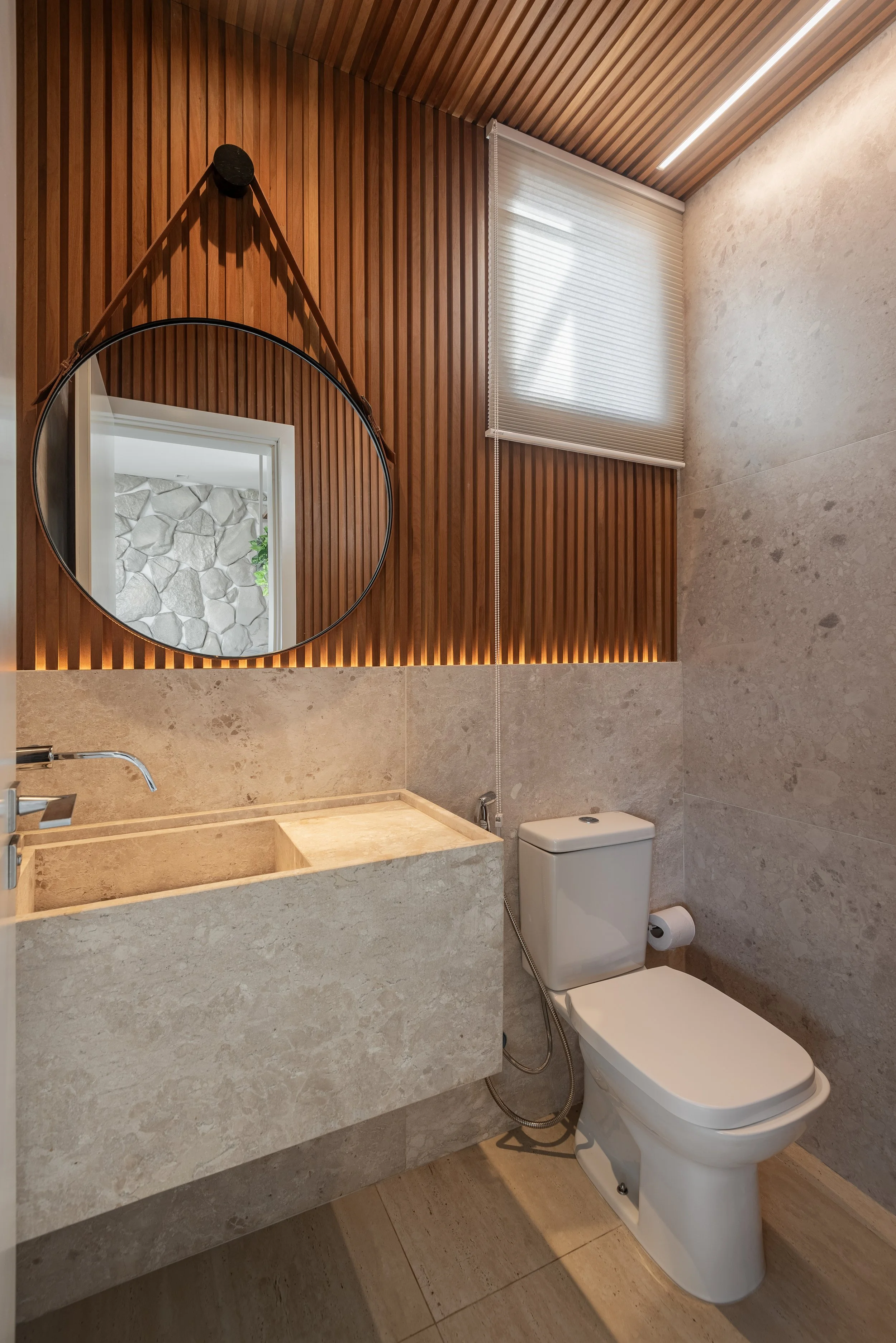 Modern bathroom featuring beige stone sink and toilet, wooden wall paneling, a round mirror, and a window with a white blind.