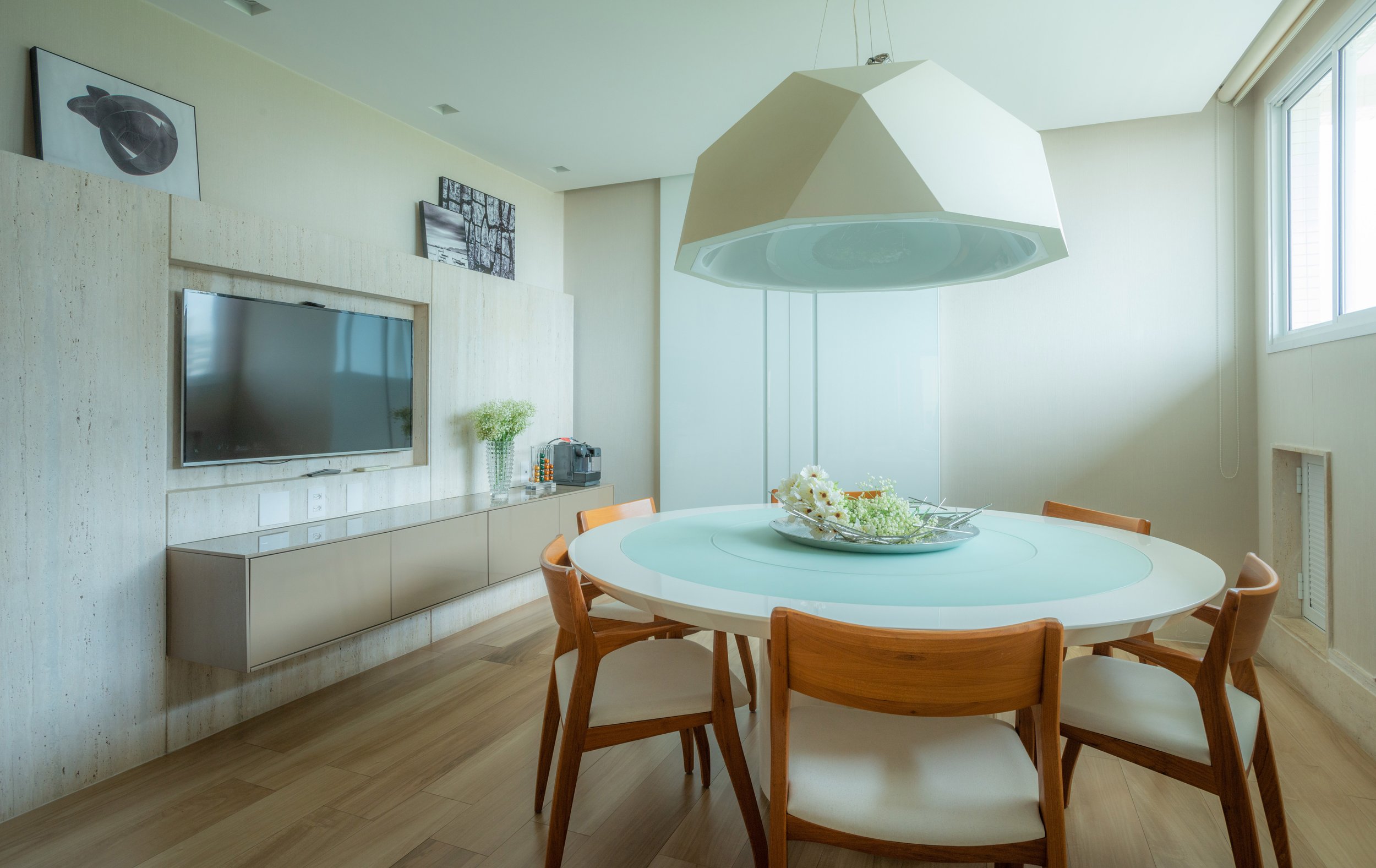 Modern dining room with a round white table, six wooden chairs, a large white pendant light, a beige built-in sideboard, wall art, a TV, a glass vase with flowers, and a window with blinds.