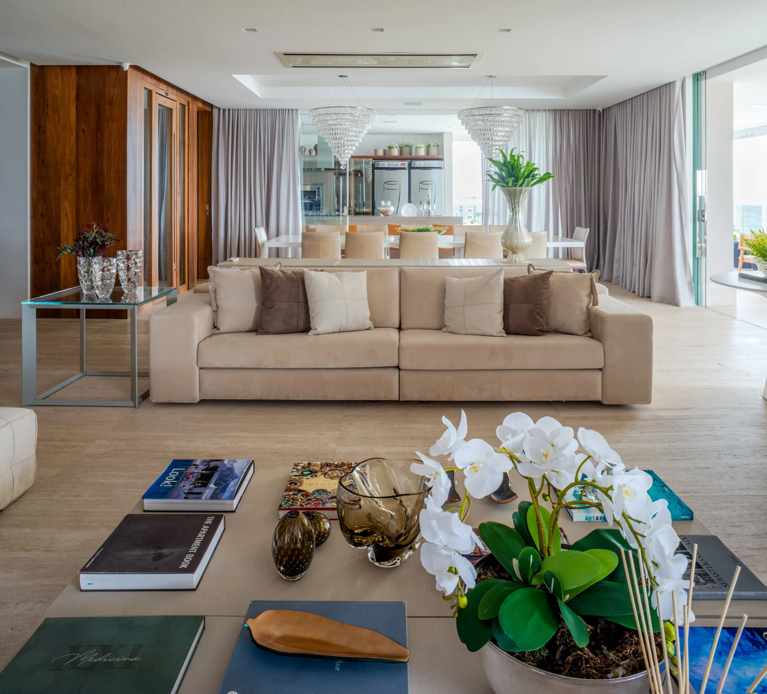 Living room with beige sofa, decorative pillows, glass table with books and decorative items, potted orchid, and dining area with large table, chairs, and chandeliers.