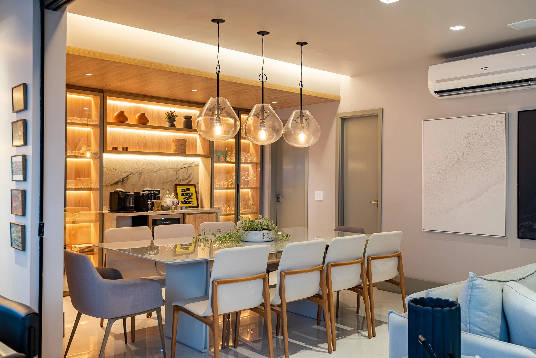 Modern dining room with a large table surrounded by white and beige chairs, hung with three glass pendant lights. A built-in wooden shelving unit with backlit shelves holding various decorative items and glassware is visible in the background. There'
