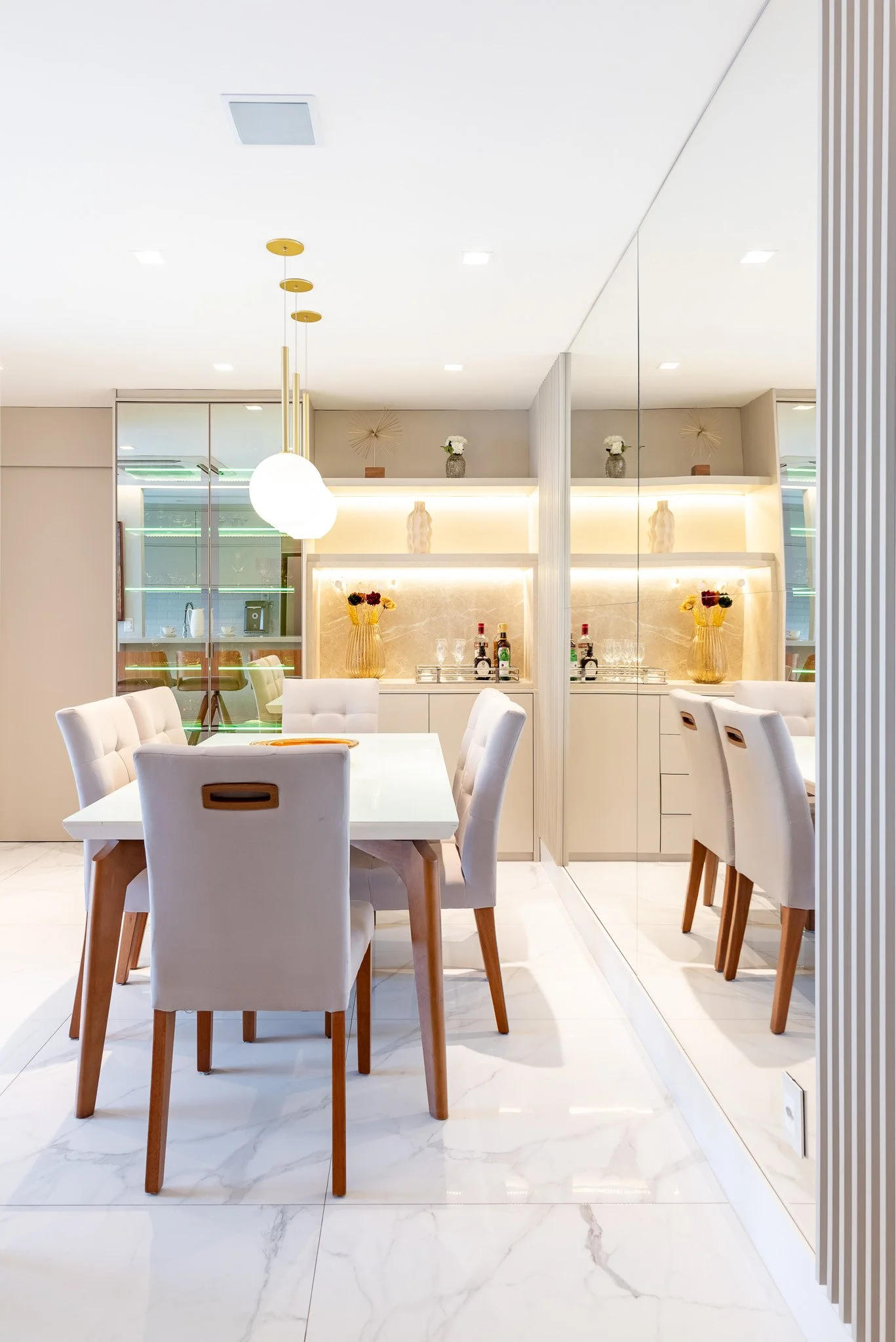 Modern dining room with white chairs around a white table, a mirror wall, and decorative vases with flowers on shelves in the background.