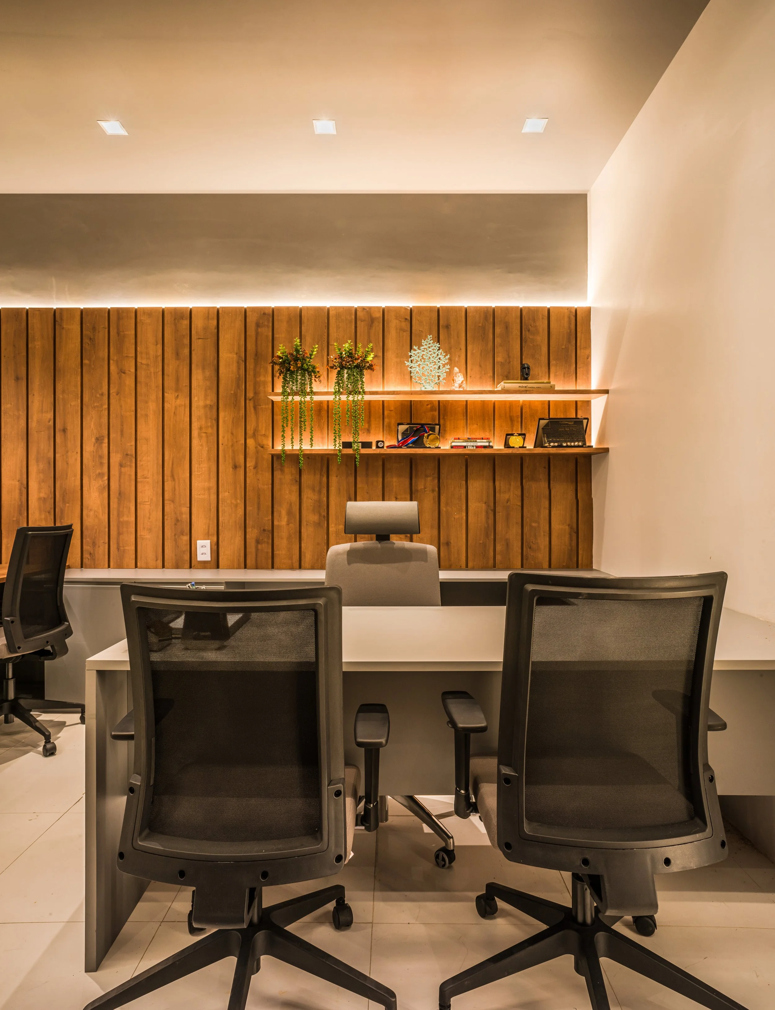 Modern office with a desk, three black office chairs, and a wooden accent wall with shelves decorated with plants, books, and decorative items.