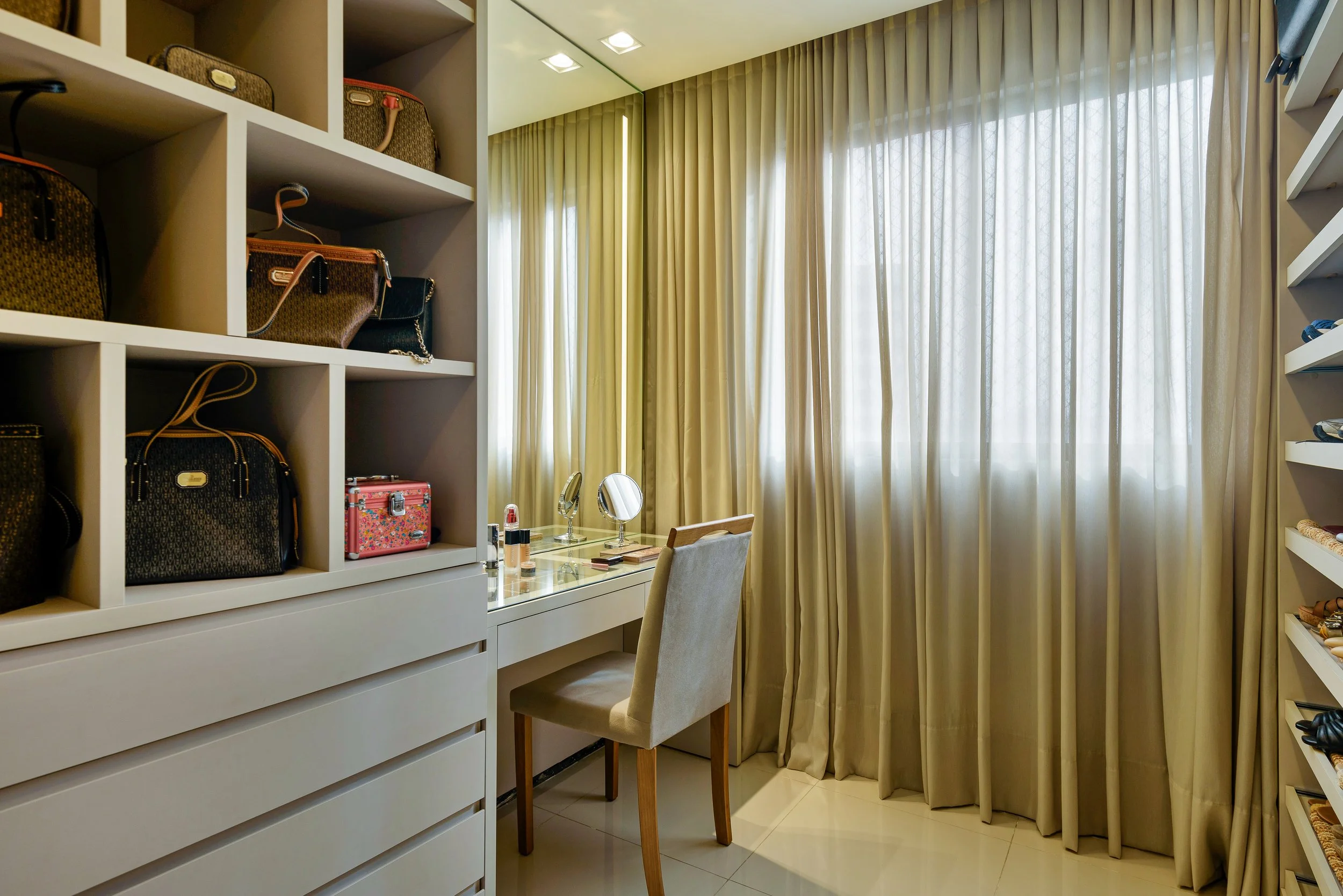 A walk-in closet with beige curtains, a white dresser, a small vanity with makeup items, and shelves with handbags and shoes.