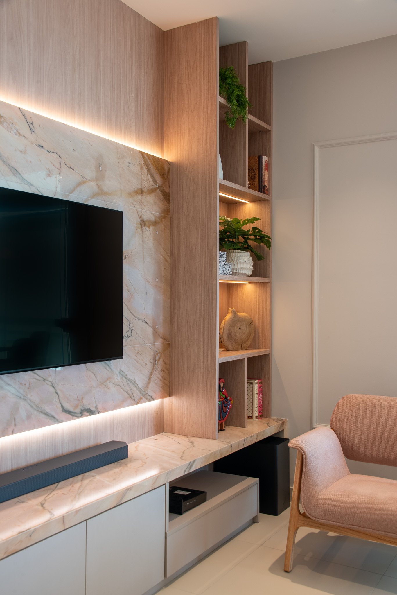 Living room with a wall-mounted flat-screen TV, marble panel behind it, and a built-in wooden bookshelf with decorative items, plants, and books, with soft lighting accents.