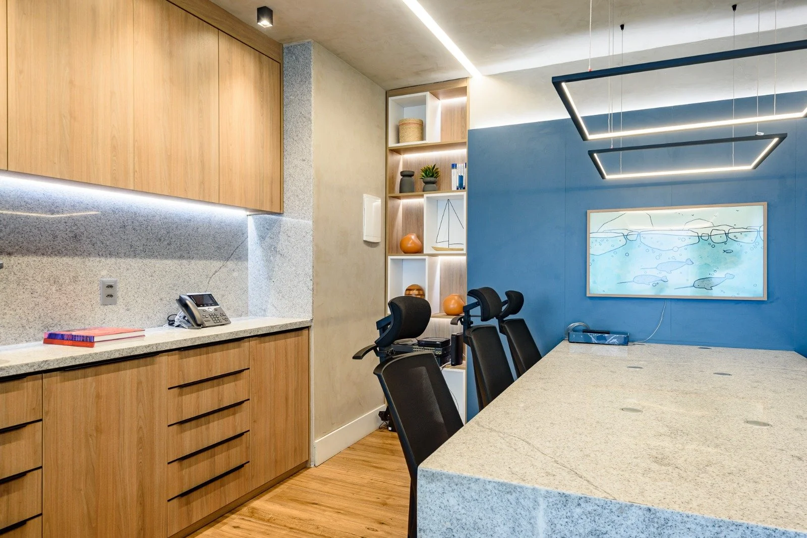 A modern conference room with a large granite table, black office chairs, a blue accent wall with a framed abstract artwork, and wooden cabinetry and shelving with decorative items.
