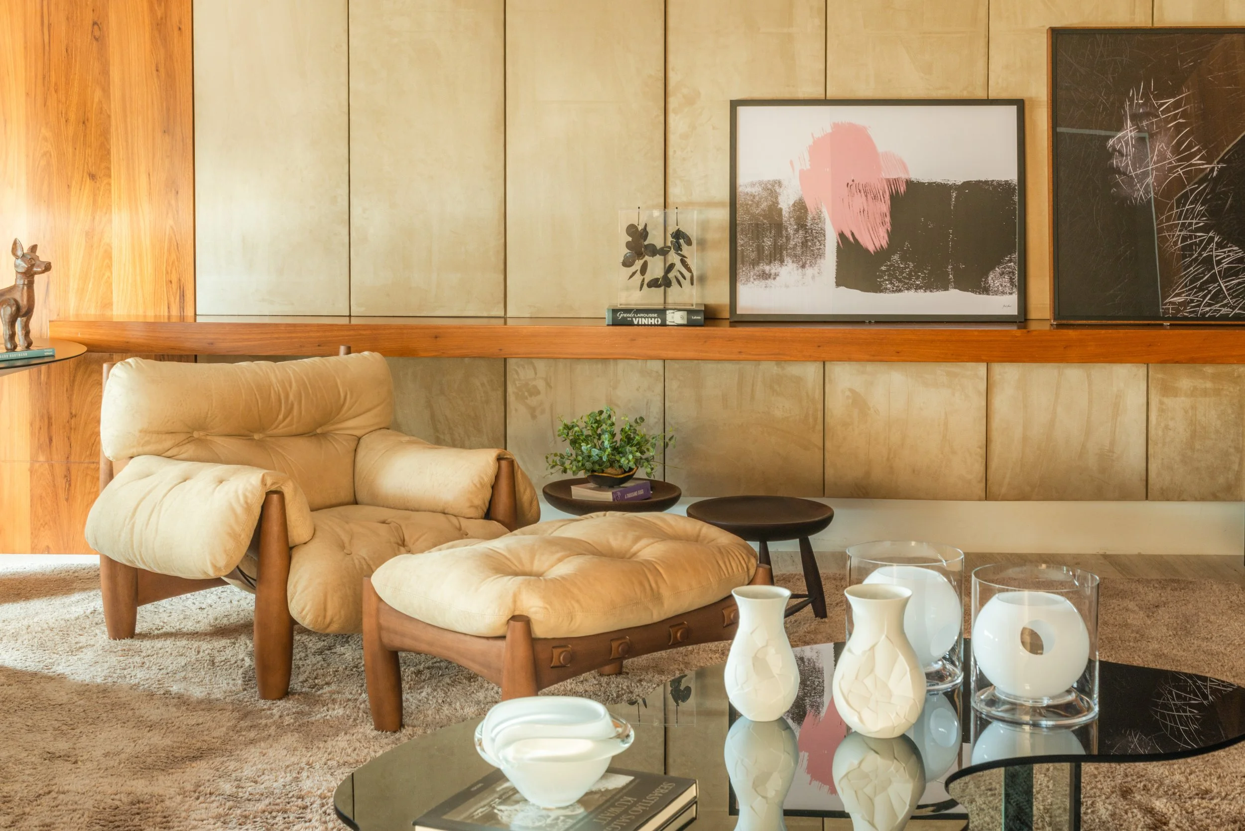 Living room with beige armchair and ottoman, wooden wall panels, abstract artwork on the wall, small coffee table with white vases, glass candle holders, and decorative objects, carpeted floor.
