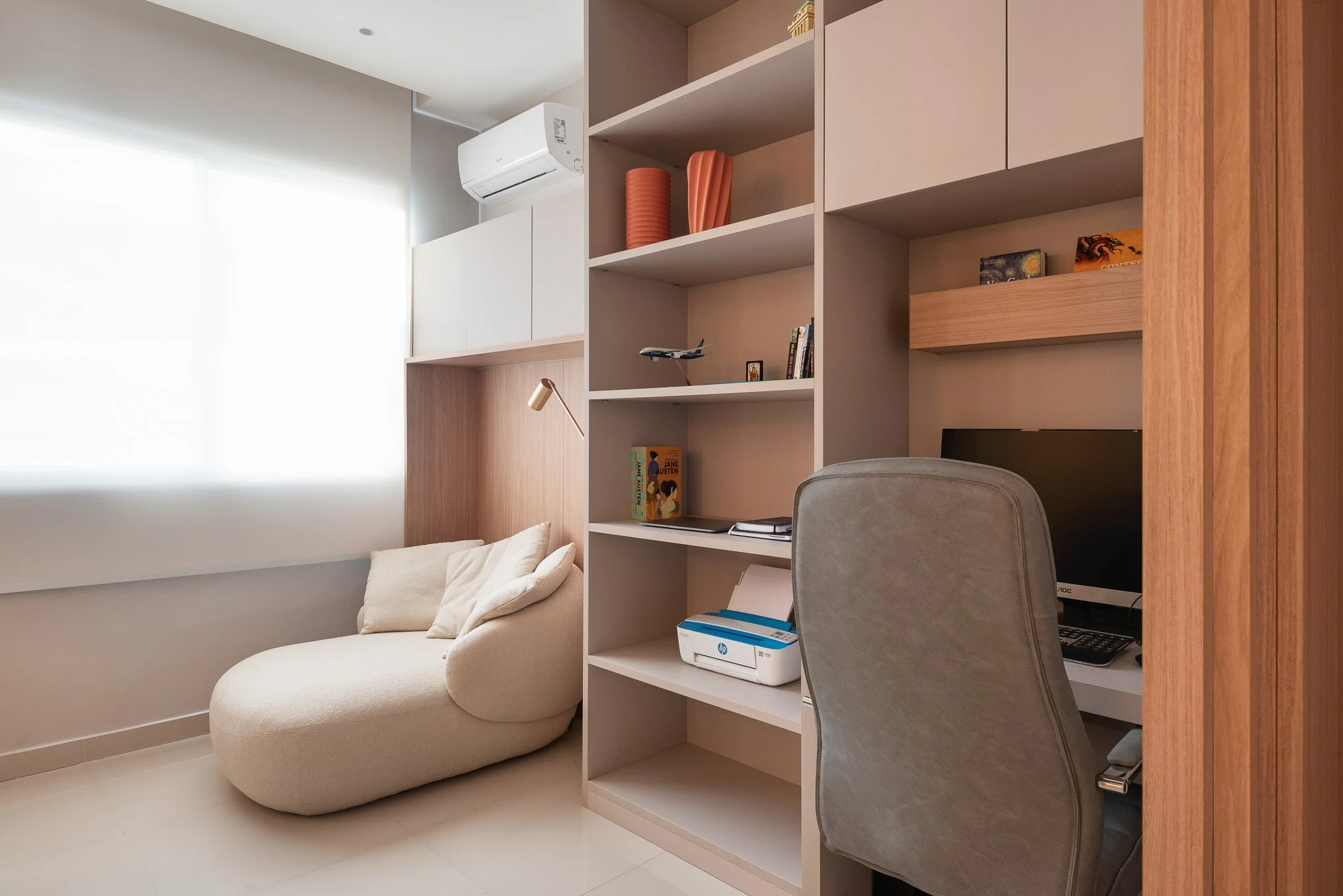 A cozy home office with a beige chair, a desk with a computer, a bookshelf with decorative items and books, and a modern lounge chair near a large window with white blinds.