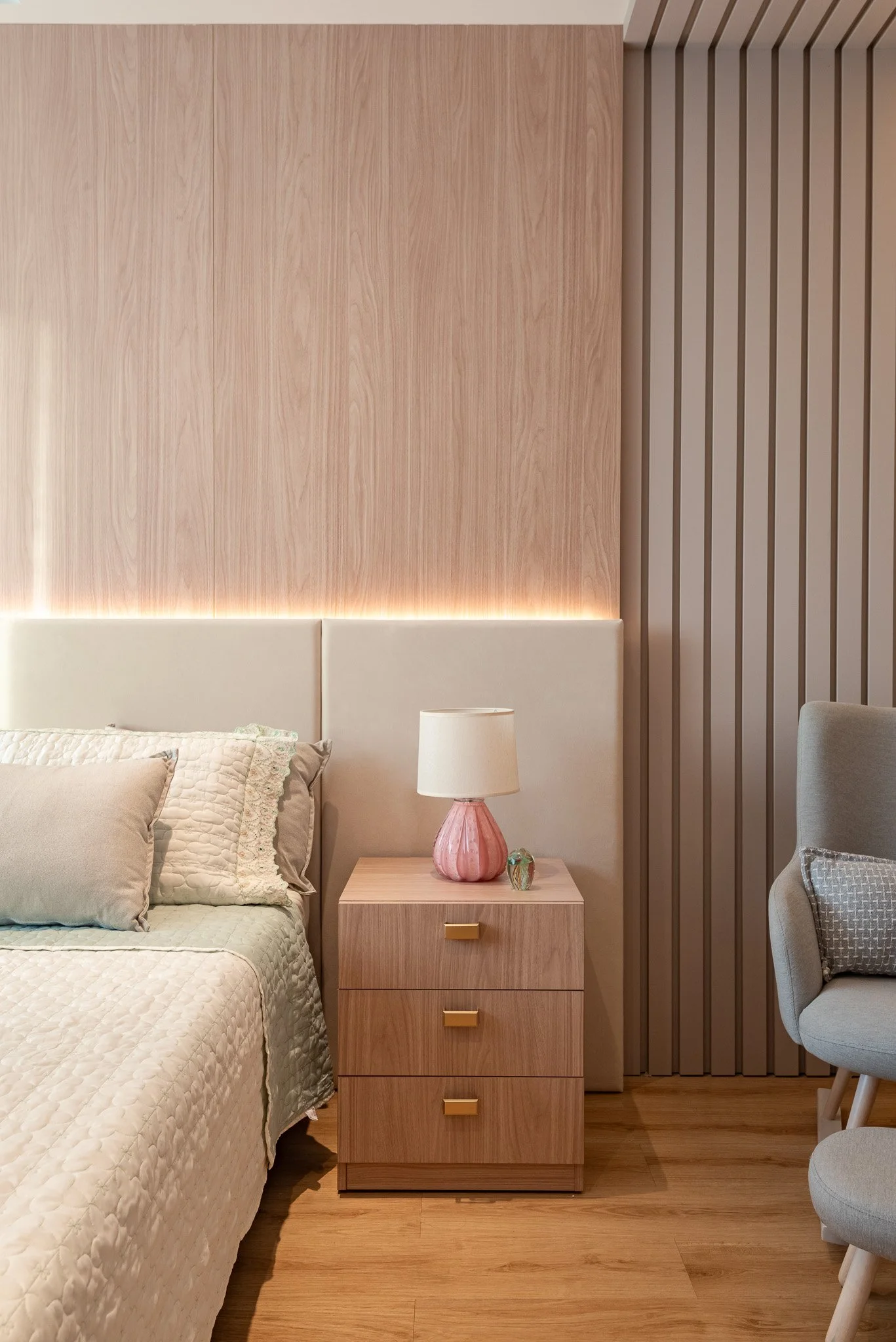 A bedroom corner with a bed featuring a quilted beige cover, two pillows, and a beige headboard. Next to the bed is a wooden nightstand with three drawers, each with a gold handle. On the nightstand is a pink vase-shaped lamp with a white shade and a