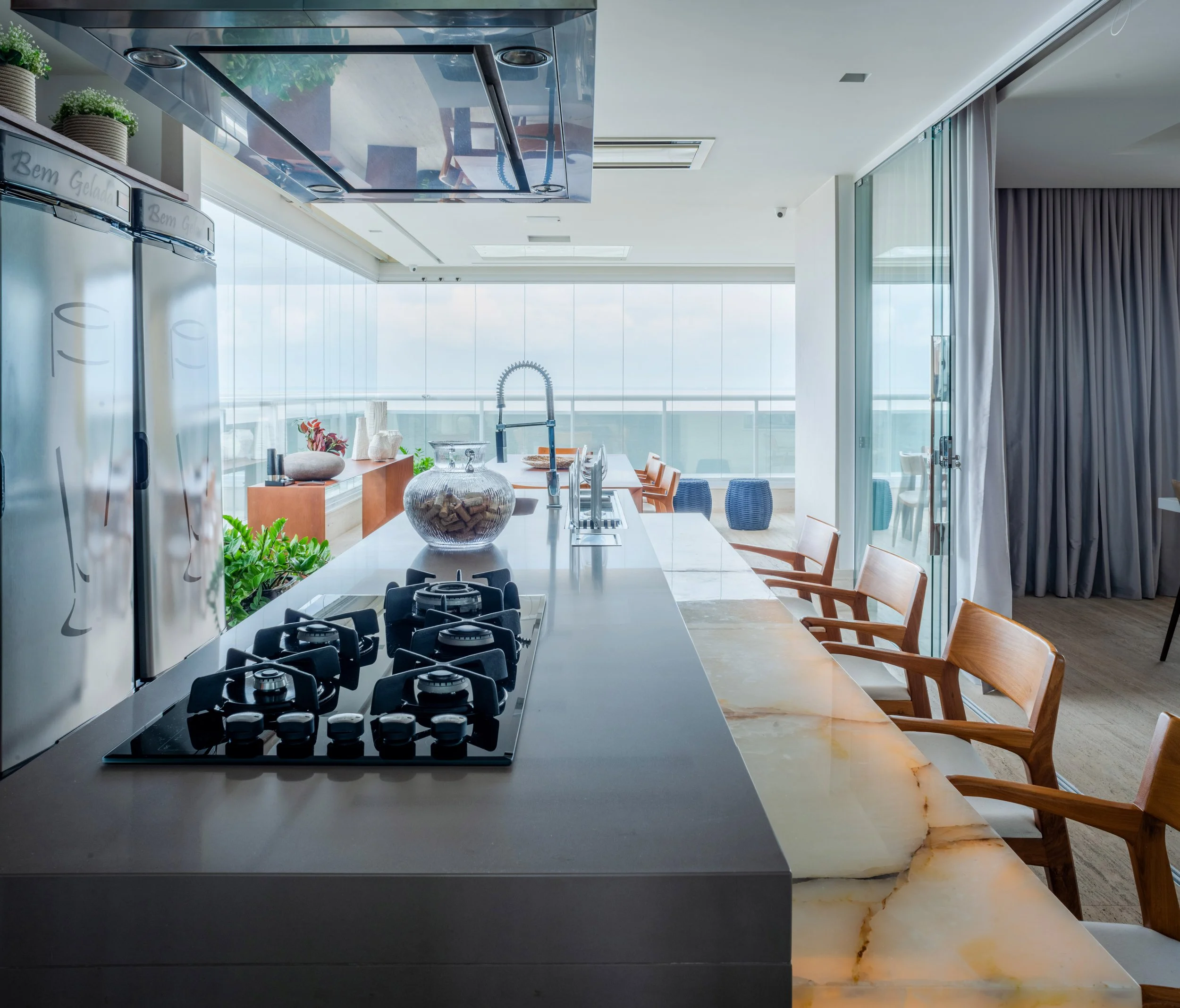 Modern kitchen with a marble countertop island, gas stove, glass jar with corks, plants, and wooden dining chairs, with a balcony view outside.