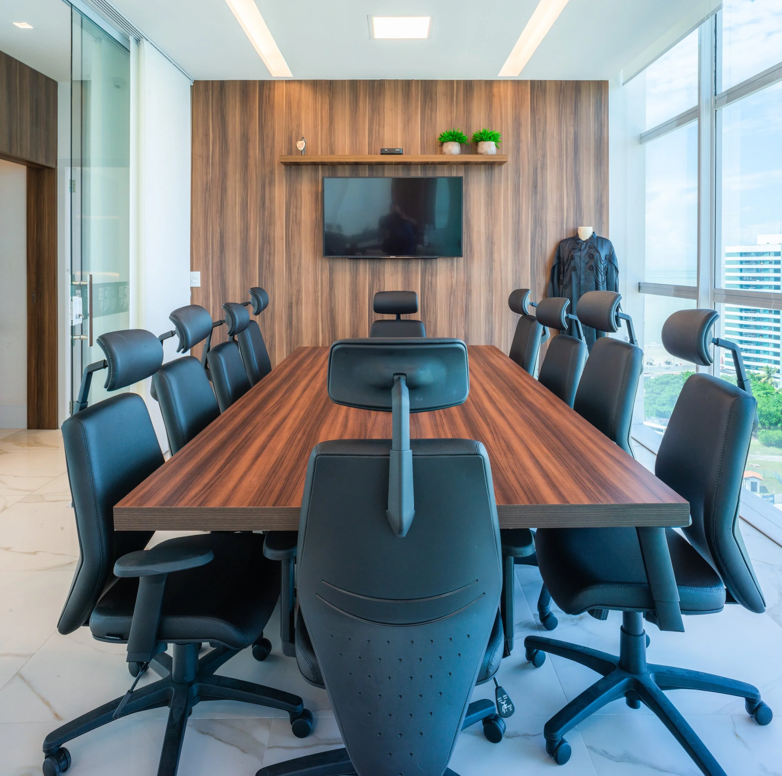 A modern conference room with a large wooden table surrounded by black office chairs. There is a large flat-screen TV on a wood-paneled wall, a small shelf with green plants, and large windows letting in natural light.