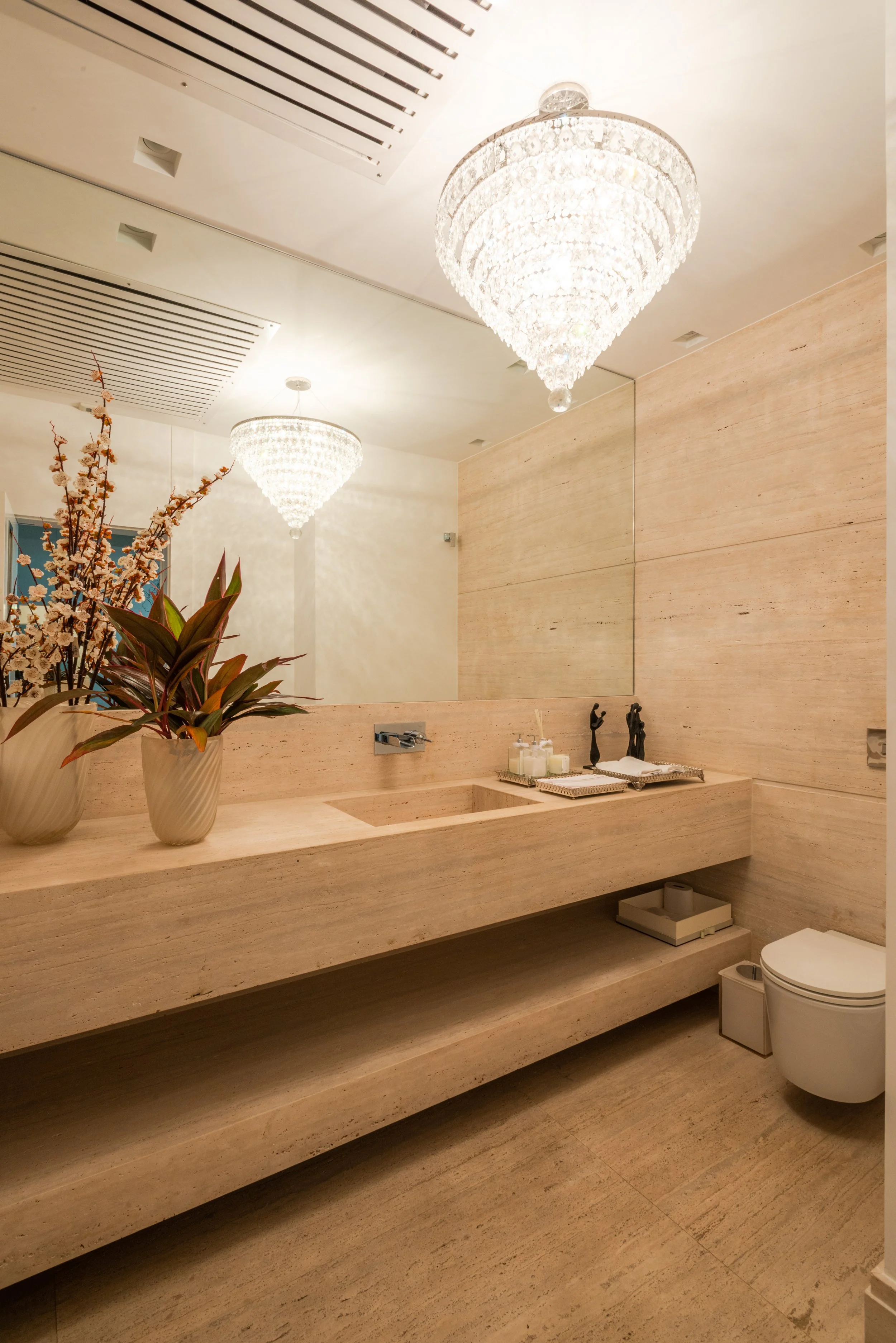 Luxurious beige bathroom with a large mirror, chandelier hanging from the ceiling, potted plants on the countertop, candles, and a modern toilet.