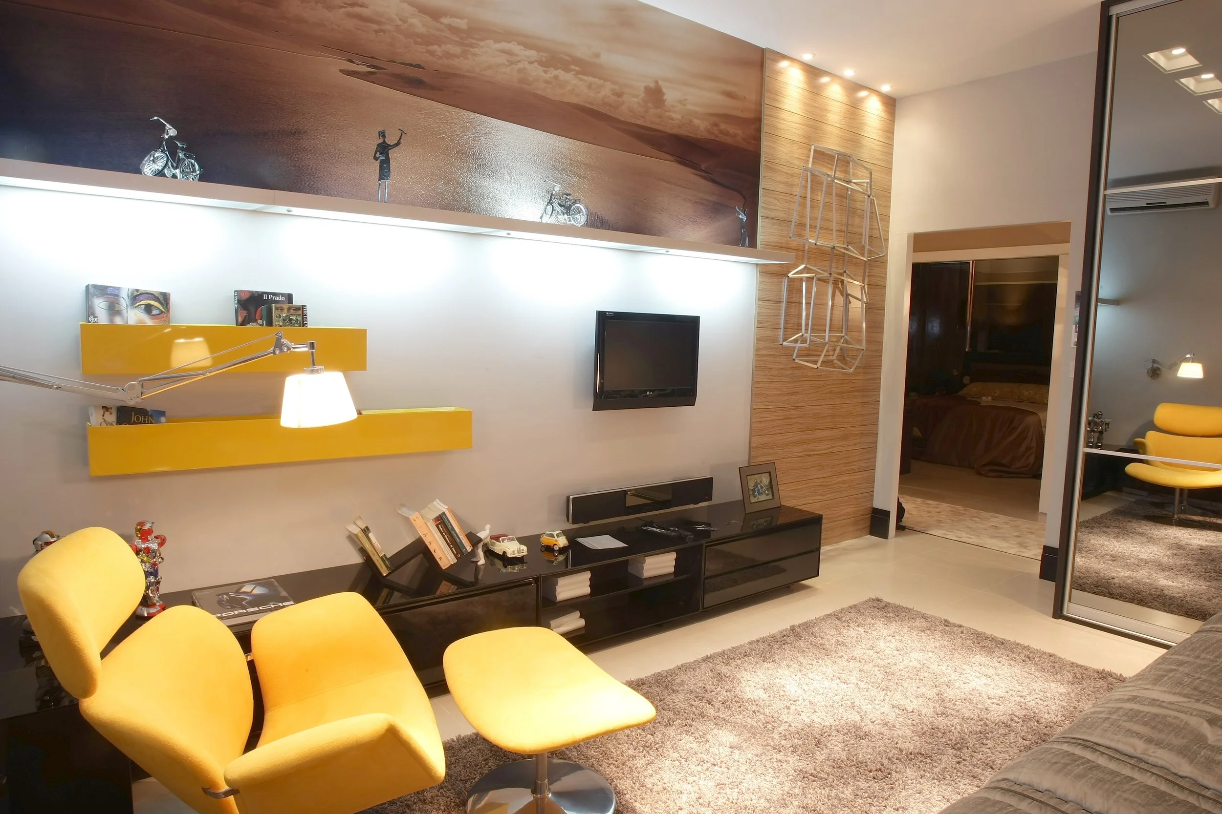 Interior of a modern living room with yellow chairs, a black entertainment center, a small TV, decorative shelves, and a mirror, with a bedroom visible through an open door.