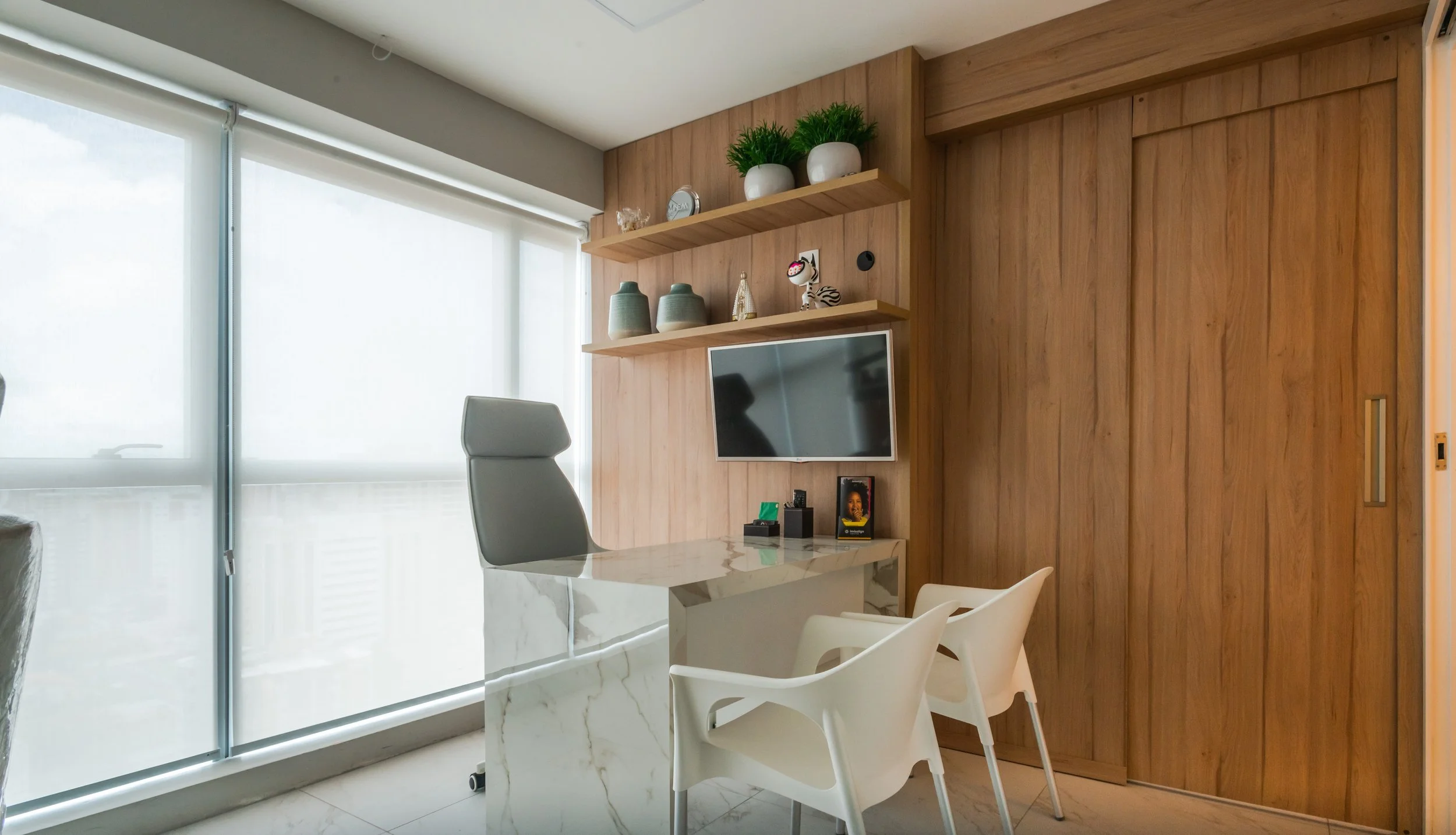 Modern office with a marble desk, gray ergonomic chair, and white chairs in front. Wooden wall with shelves holding decorative items, a small TV, and a sliding wooden door.