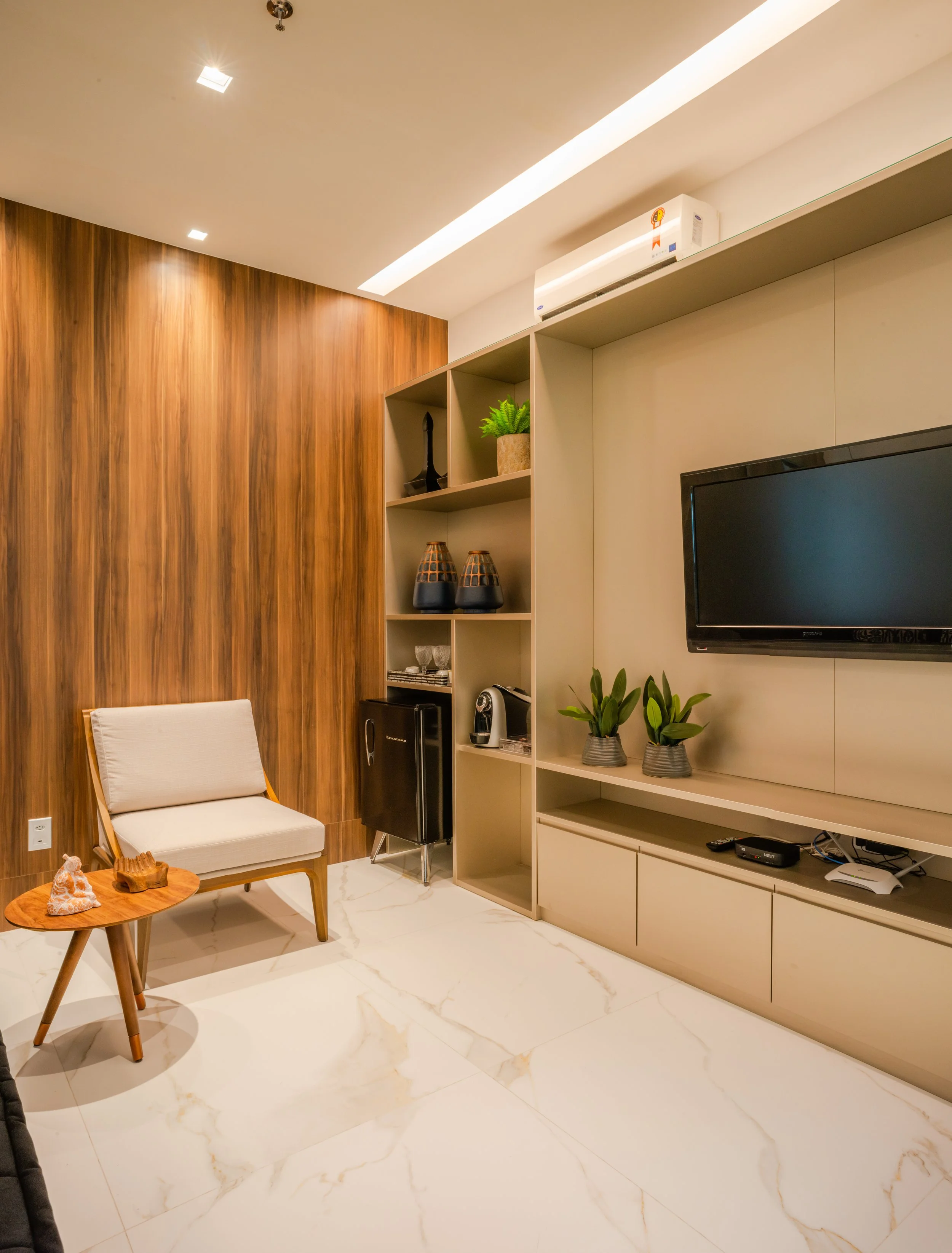Modern living room with a beige armchair, a small wooden side table, potted plants, mounted TV, and decorative items on white shelving against a wood-paneled wall, with marble flooring.
