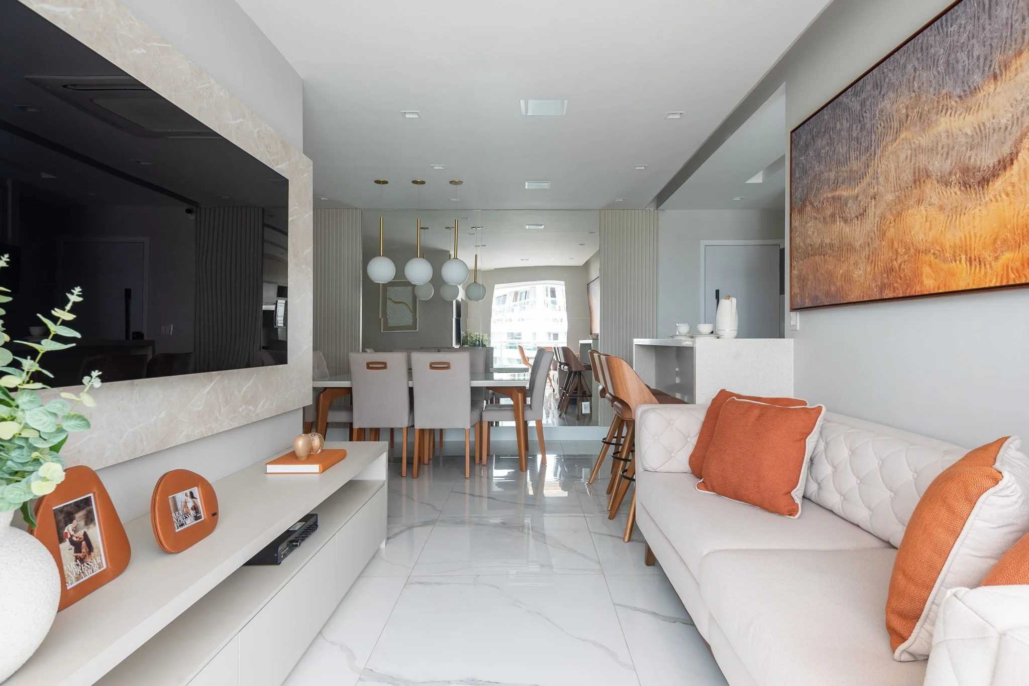 Modern living room with white sofa, orange pillows, and dining area with white chairs and hanging globe lights, in a bright apartment.
