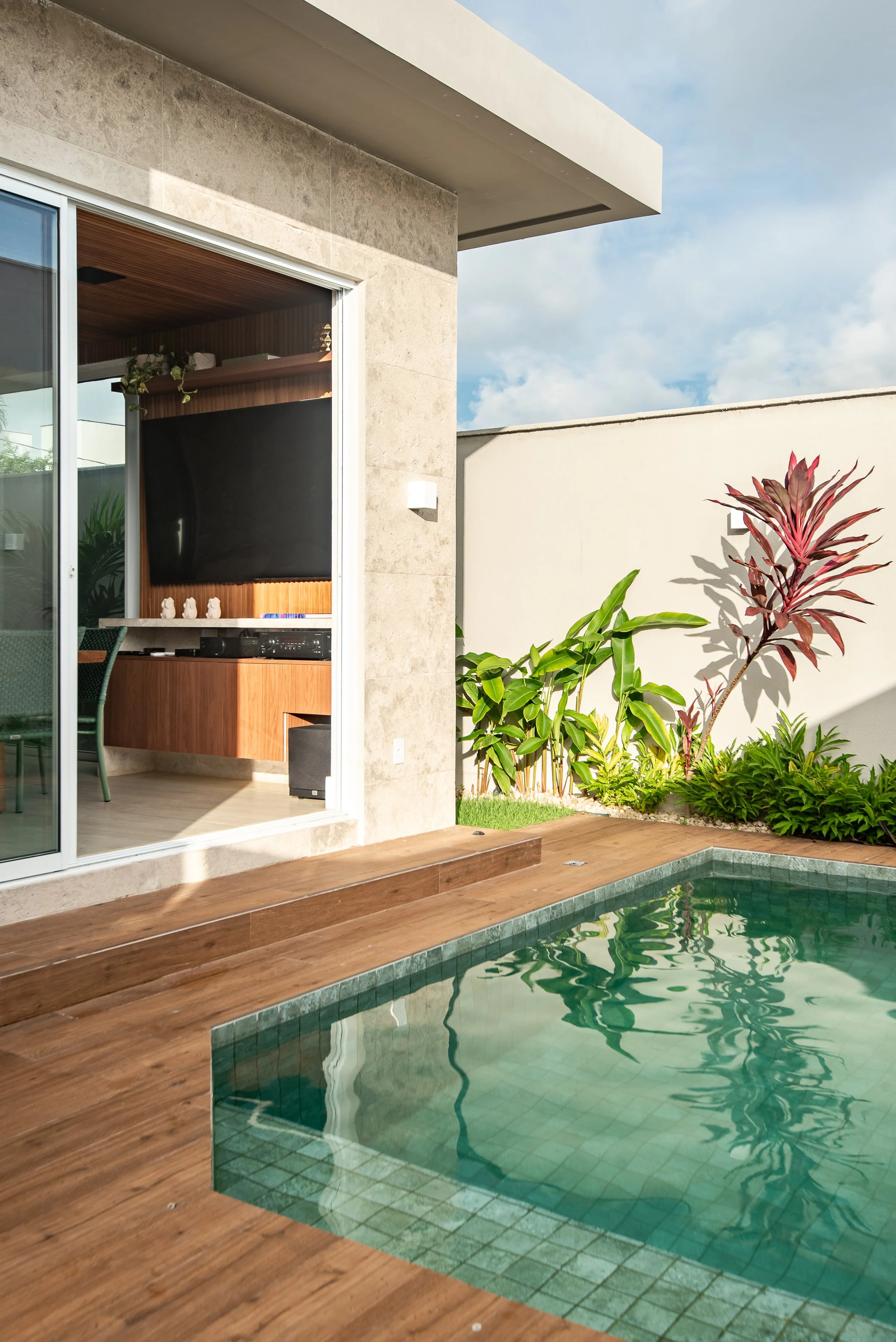 A modern backyard with a small swimming pool, wooden deck, sliding glass door, and lush tropical plants against a white wall.
