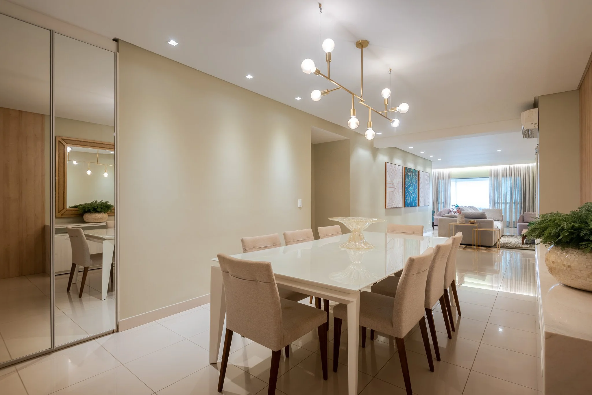 Modern dining room with a large white table, beige upholstered chairs, and a glass bowl centerpiece. The room has a gold and white chandelier, light-colored walls, and tiled floor. In the background, there is a living area with sofas and artwork on t