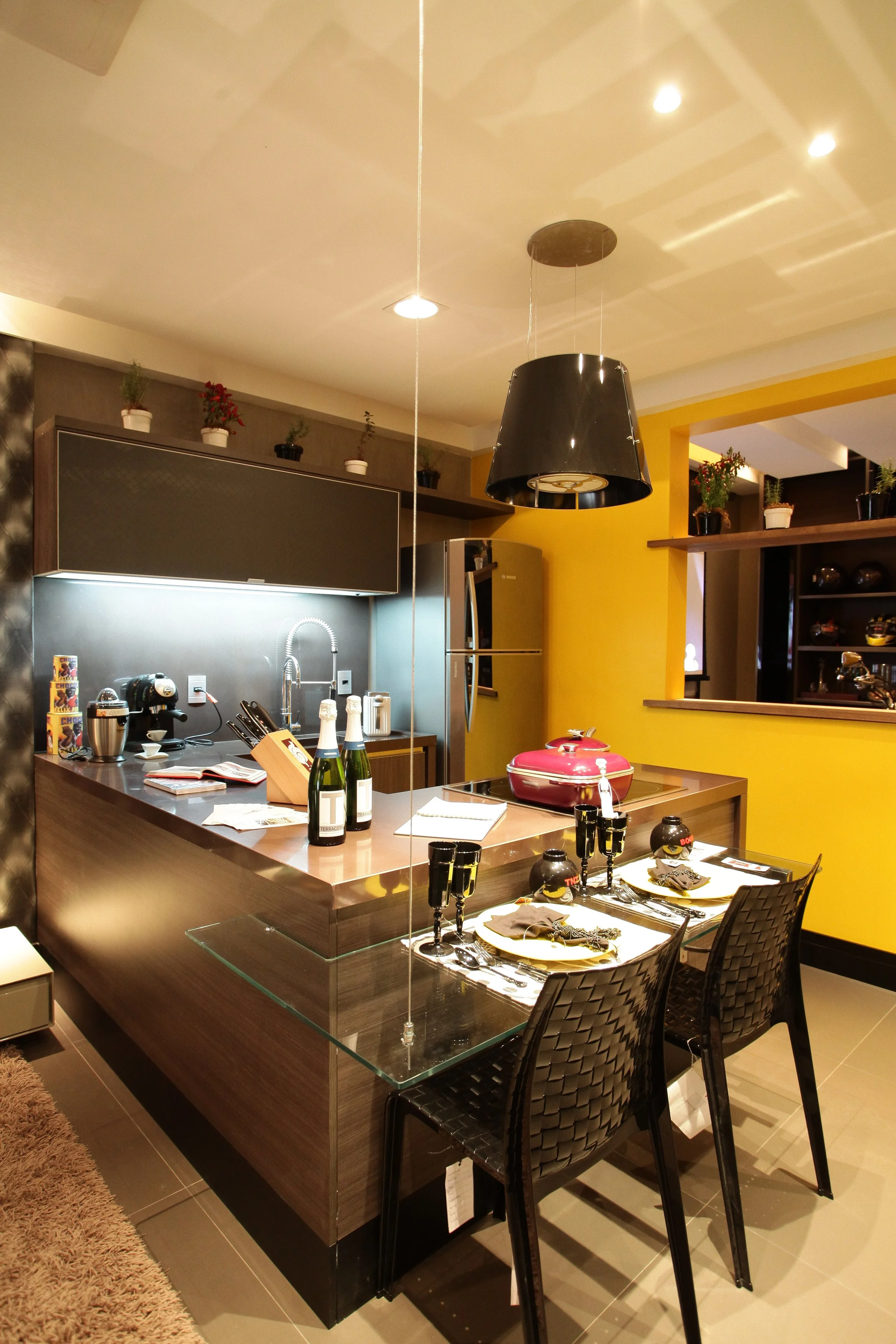 A modern kitchen with yellow and brown walls, a central island with a pink container, set dining table with black chairs, three champagne bottles, black glasses, and tableware, stainless steel appliances, small potted plants, and decorative shelves w