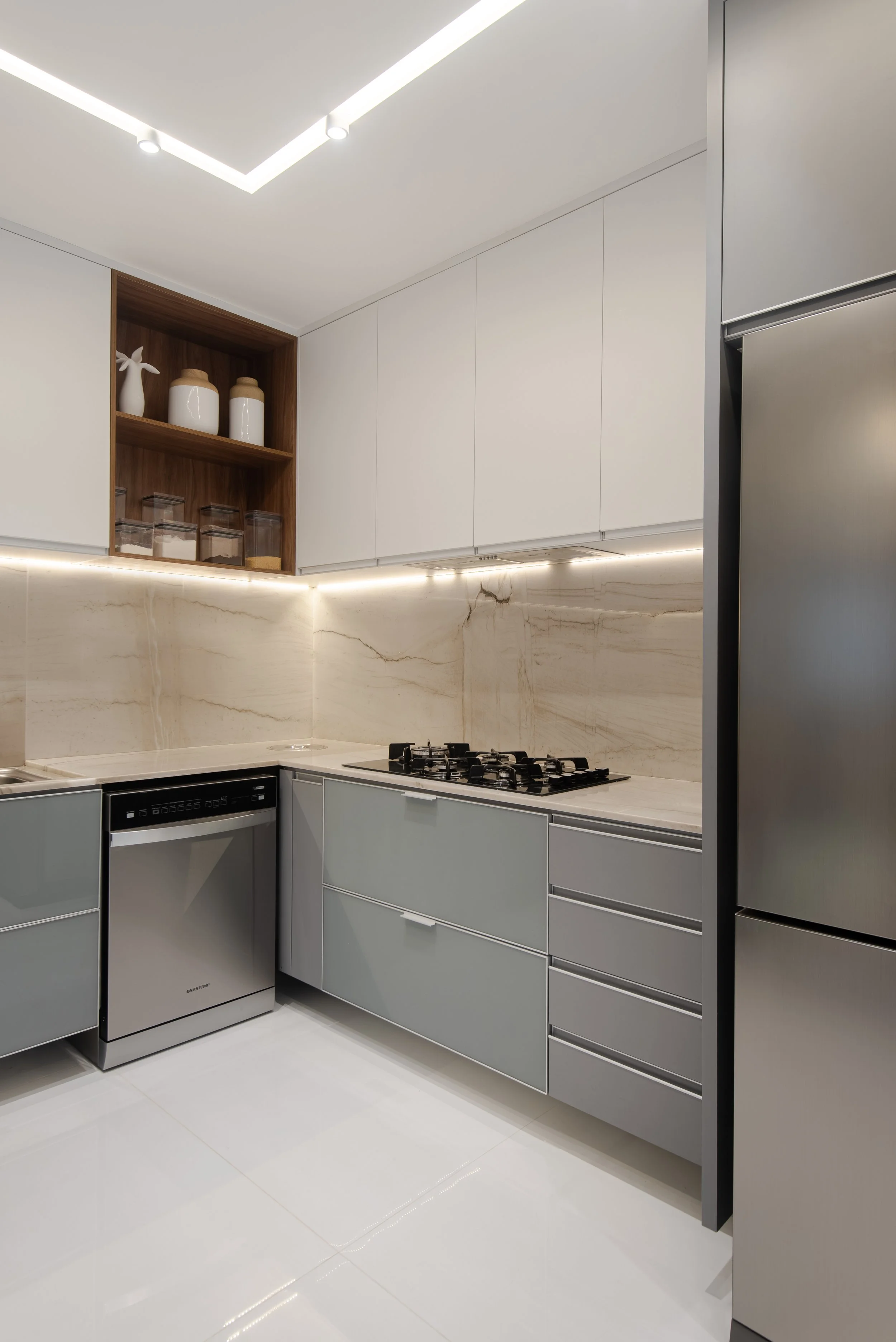 Modern kitchen with gray cabinets, marble backsplash, stainless steel refrigerator, dishwasher, and a four-burner gas stove, with open shelves holding decorative jars and vases.