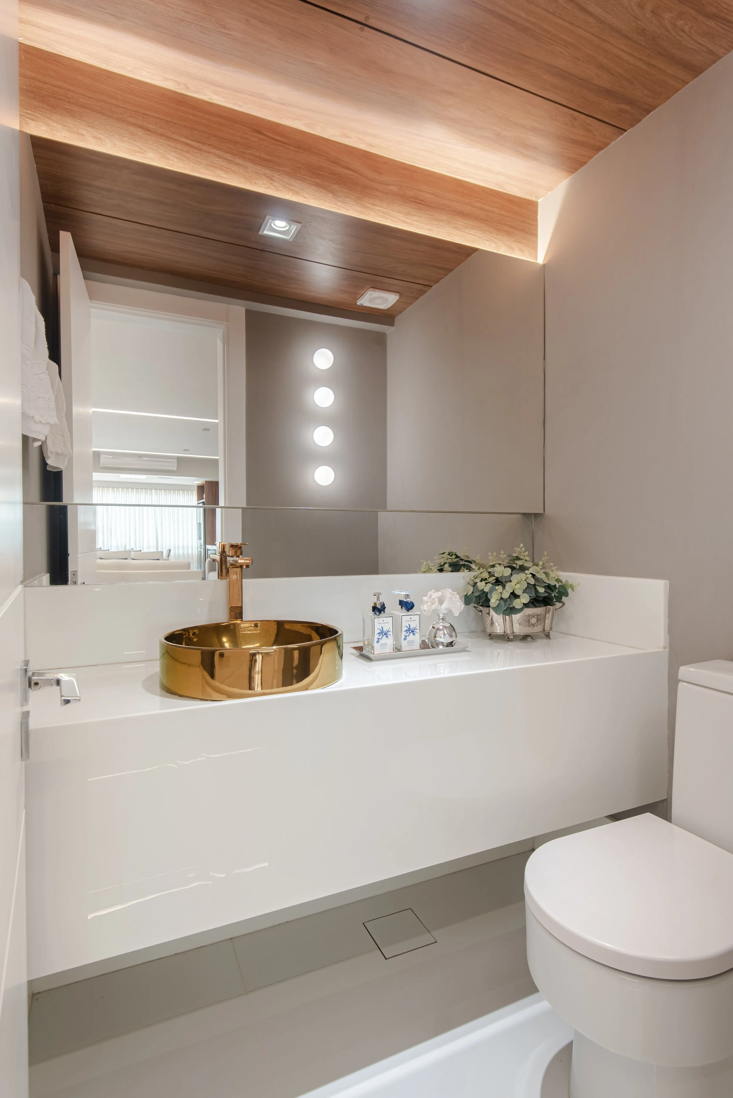 Modern bathroom with a white vanity, gold sink, decorative plant, soap bottles, and minimal decor, with wooden ceiling and soft lighting.