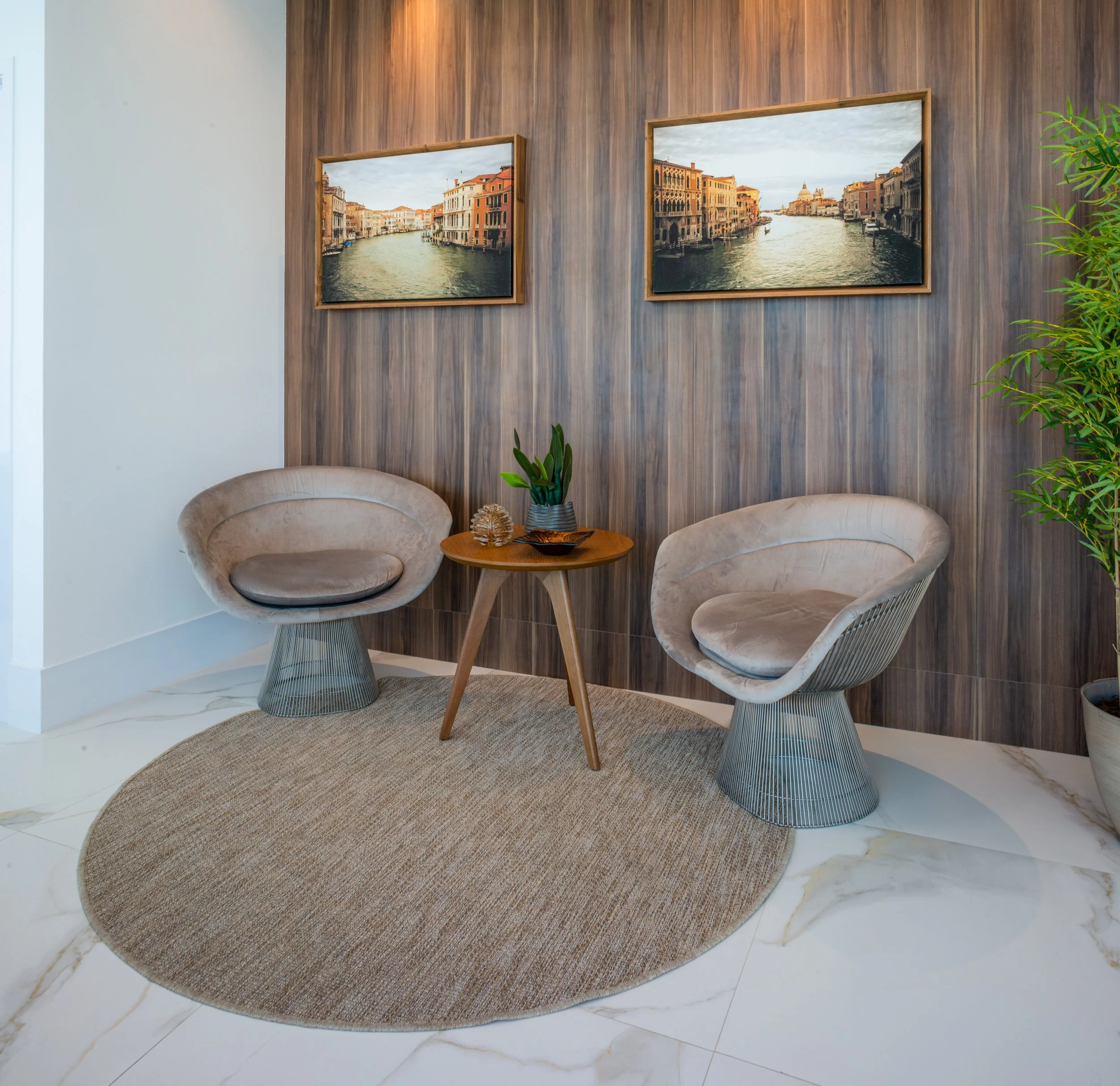 A cozy sitting area with two modern beige armchairs, a small wooden table with a potted plant, set on a round beige rug, against a wood-paneled wall decorated with two framed pictures of Venice canals.
