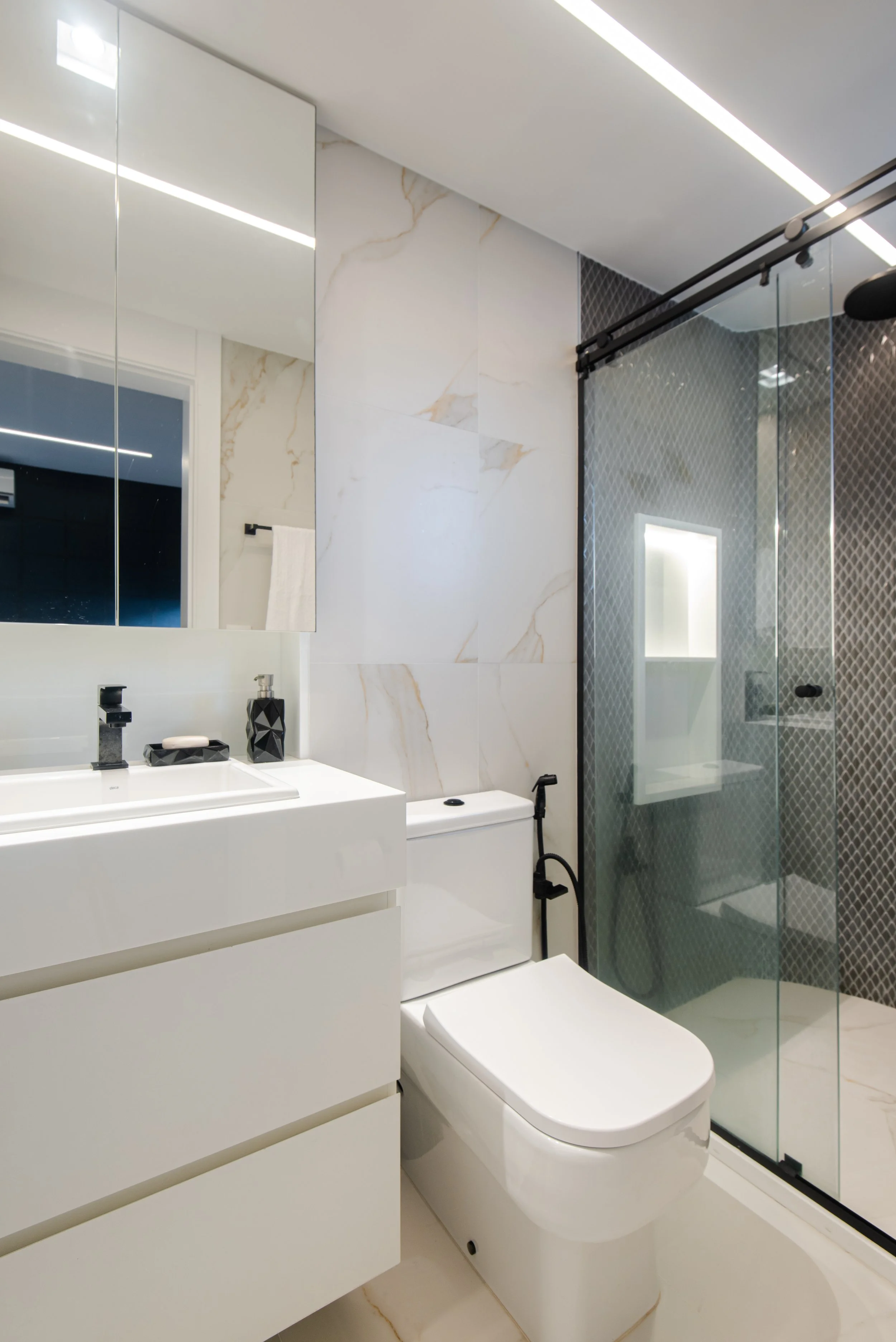 Modern bathroom with white vanity, black faucet, black soap dispenser, wall-mounted mirror, and a glass-enclosed shower with textured dark tiles.