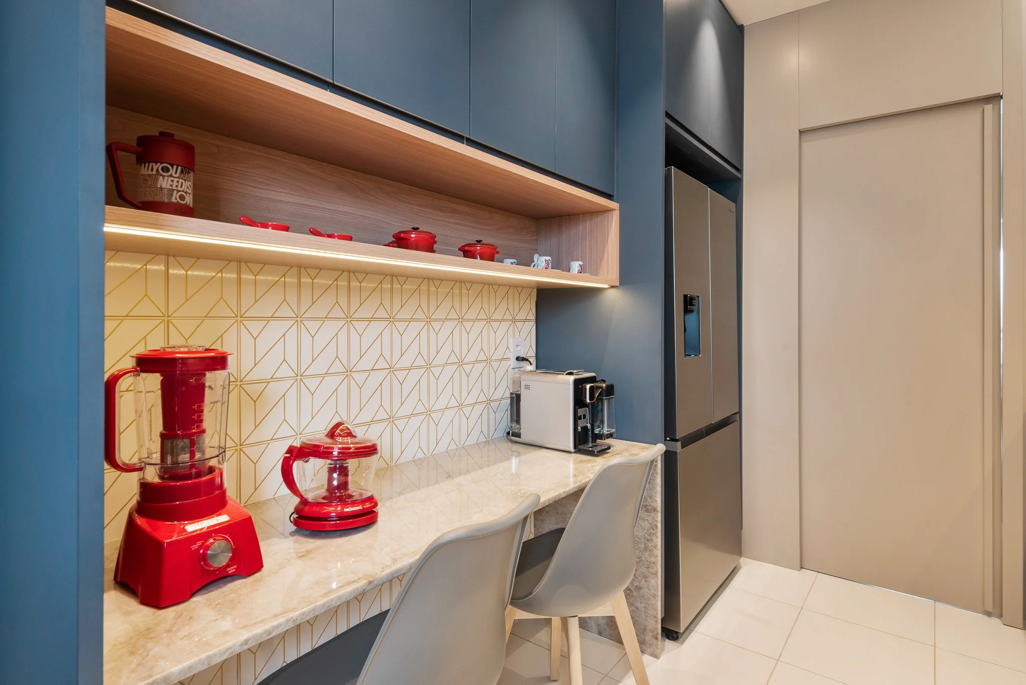 Modern kitchen with beige countertop, blue and beige cabinets, yellow geometric tile backsplash, red appliances, coffee maker, and chairs.