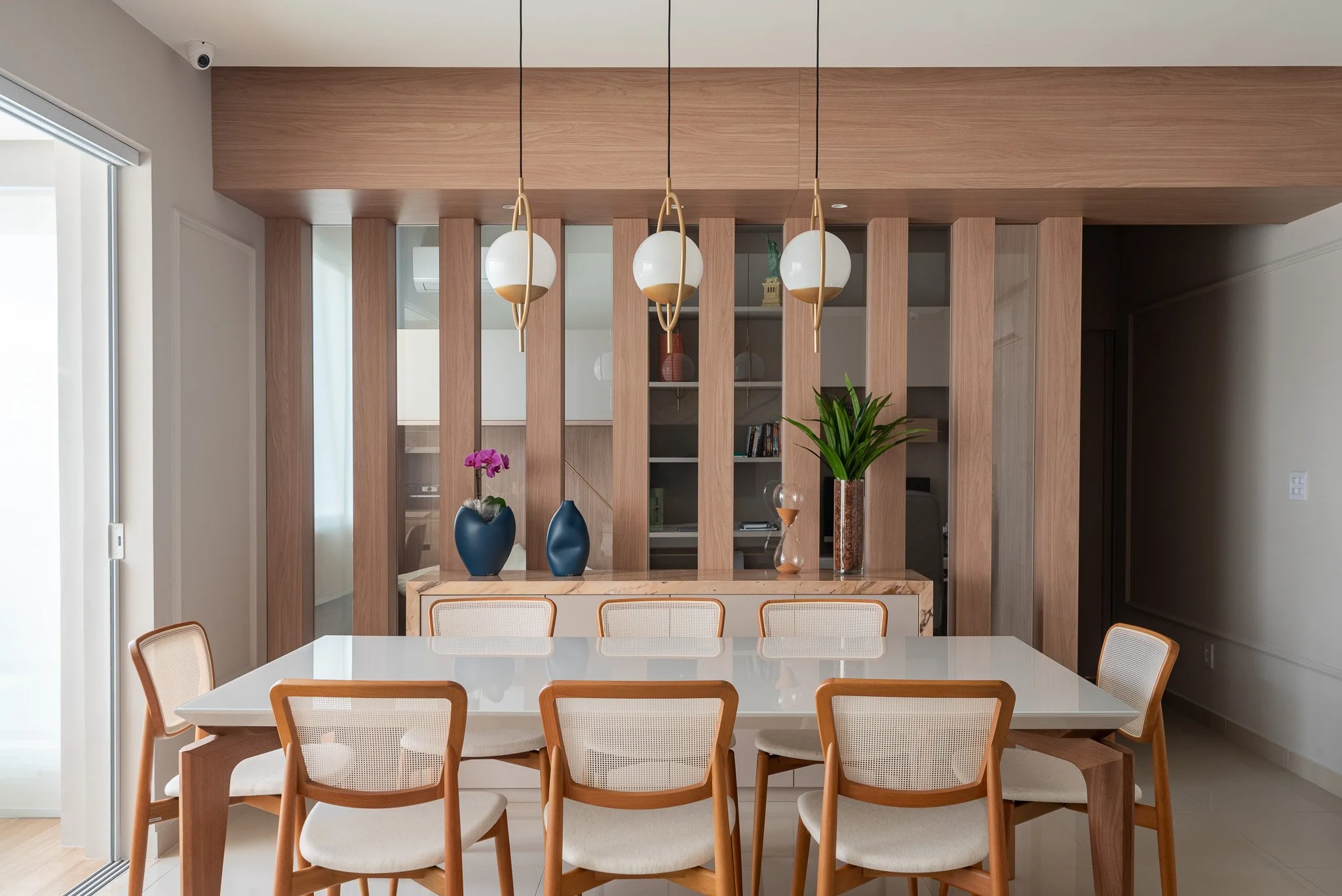 Modern dining room with a white table, wooden chairs with beige cushions, and a wooden slatted wall with built-in shelves, decorated with vases and plants. Hanging pendant lights above the table.