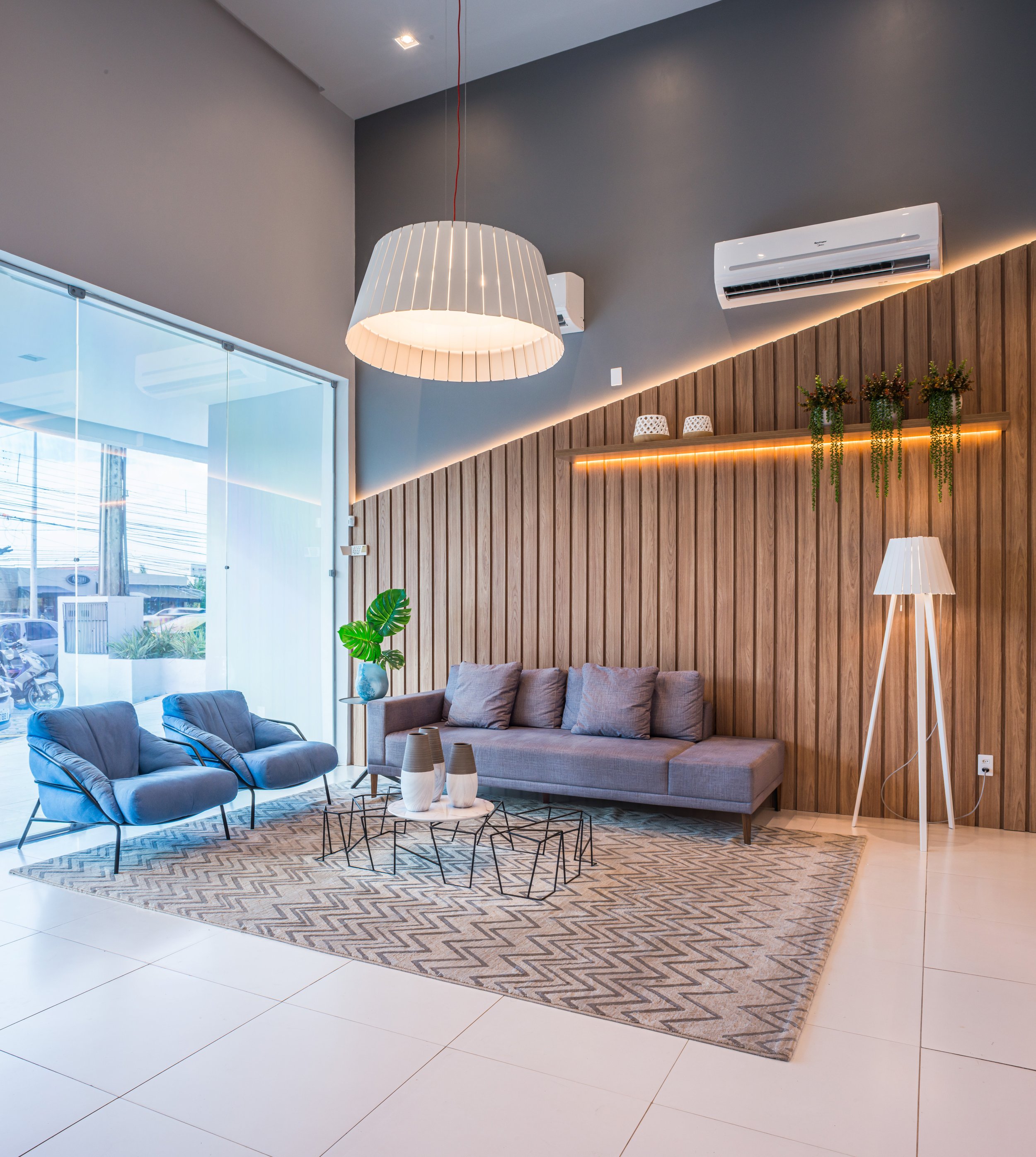 Modern hotel lobby with gray sofa, two blue armchairs, a geometric patterned rug, a white floor lamp, and a glass wall leading outside. Wooden wall panel with hanging greenery, decorative vases, and ambient lighting.