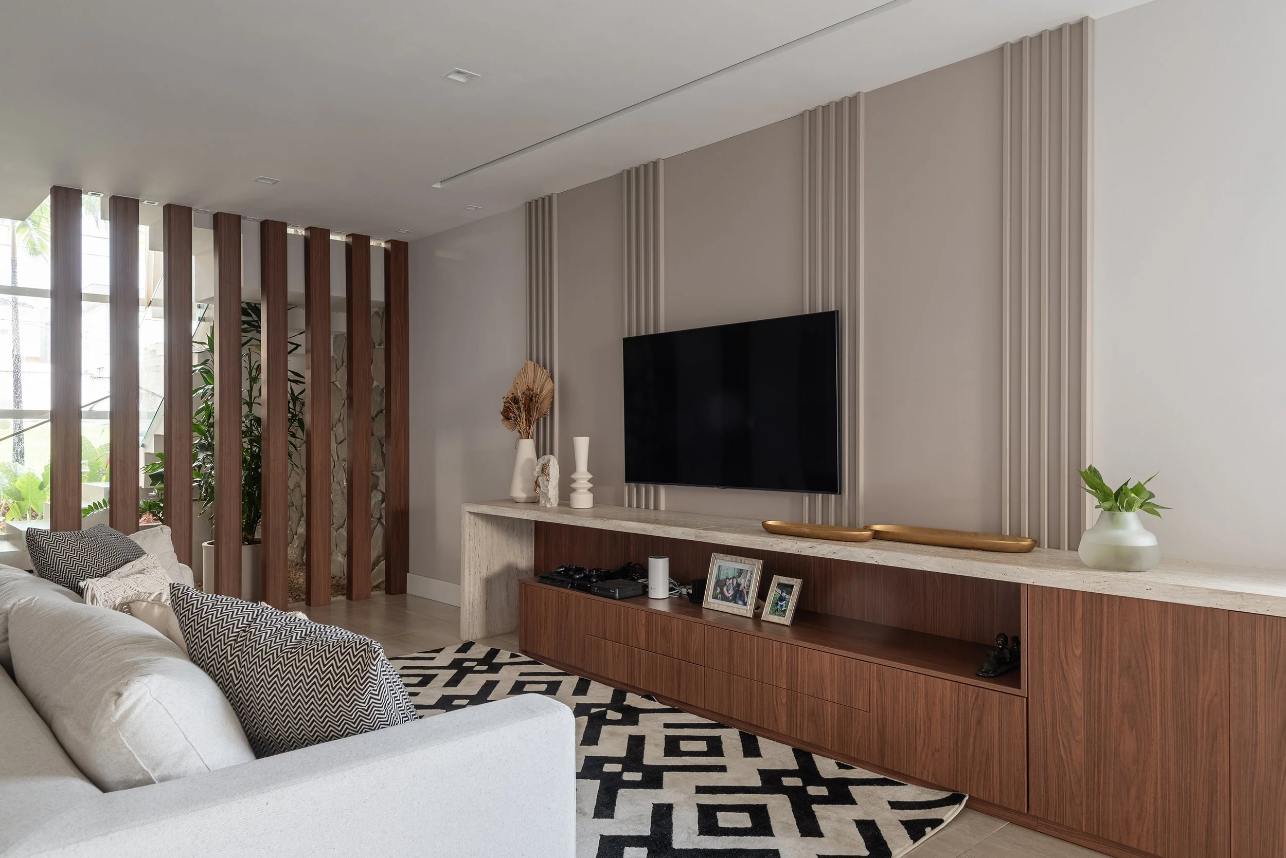 Modern living room with white sofa, patterned pillows, wooden and marble entertainment console, wall-mounted flat-screen TV, decorative vases, framed photos, black and white geometric rug, and a wooden slat room divider near a window with greenery ou