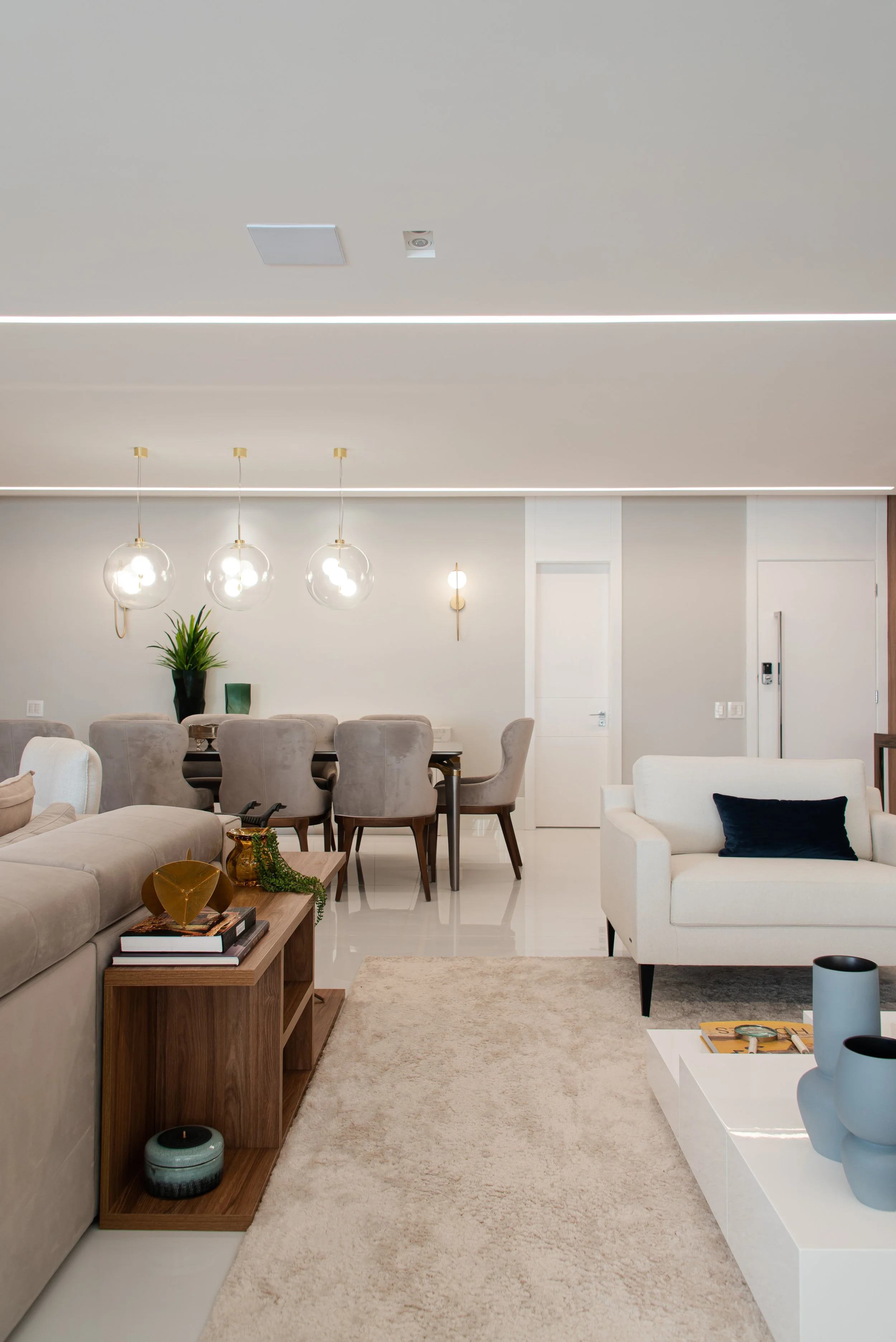 Modern living room with white walls, beige sofa, white armchair with black pillow, wooden coffee table with vases, beige rug, and a dining area with gray chairs and glass globes light fixtures.