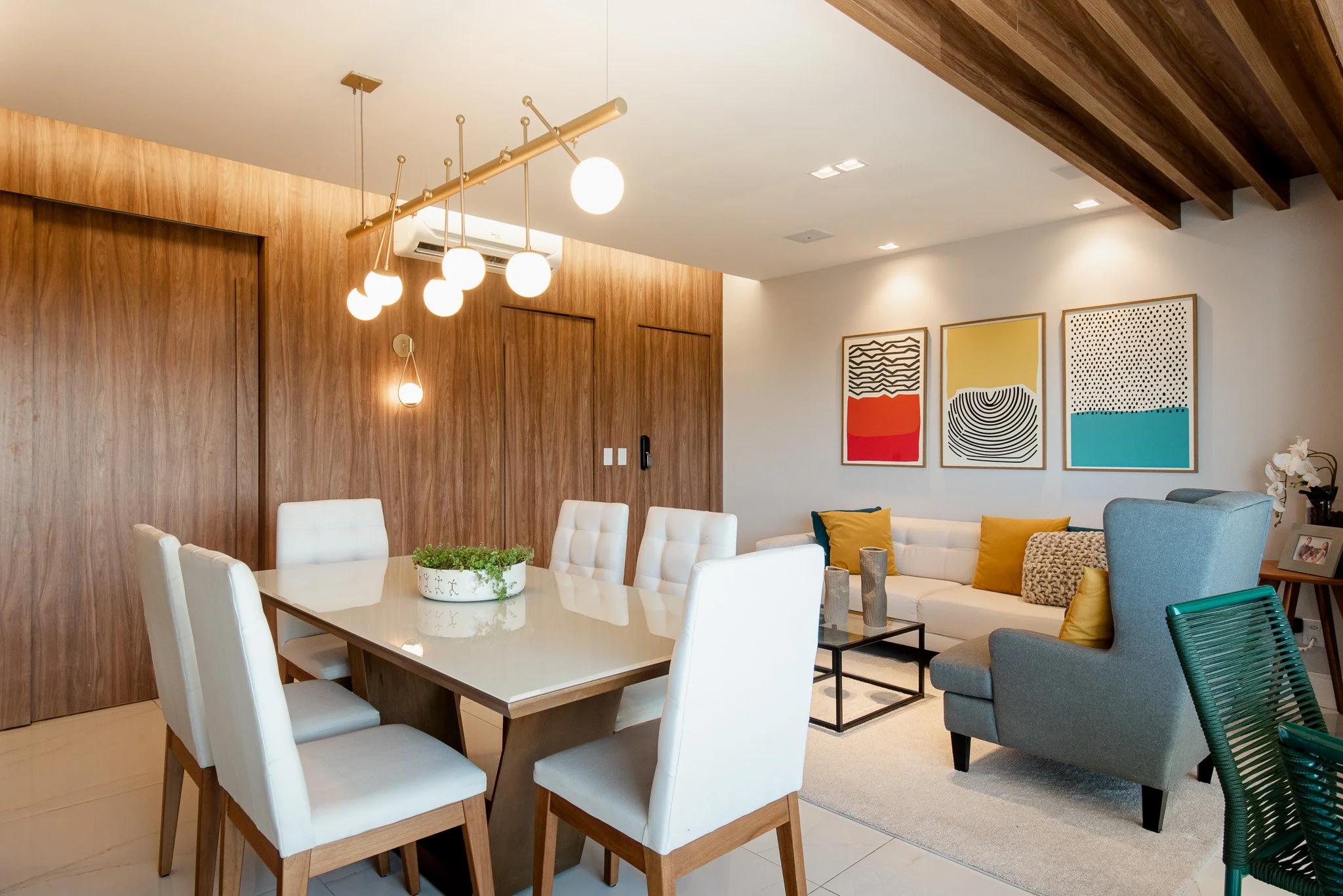 Modern living room and dining area with white dining table, six white chairs, a beige couch with colorful pillows, abstract artwork on the wall, and wooden accents.