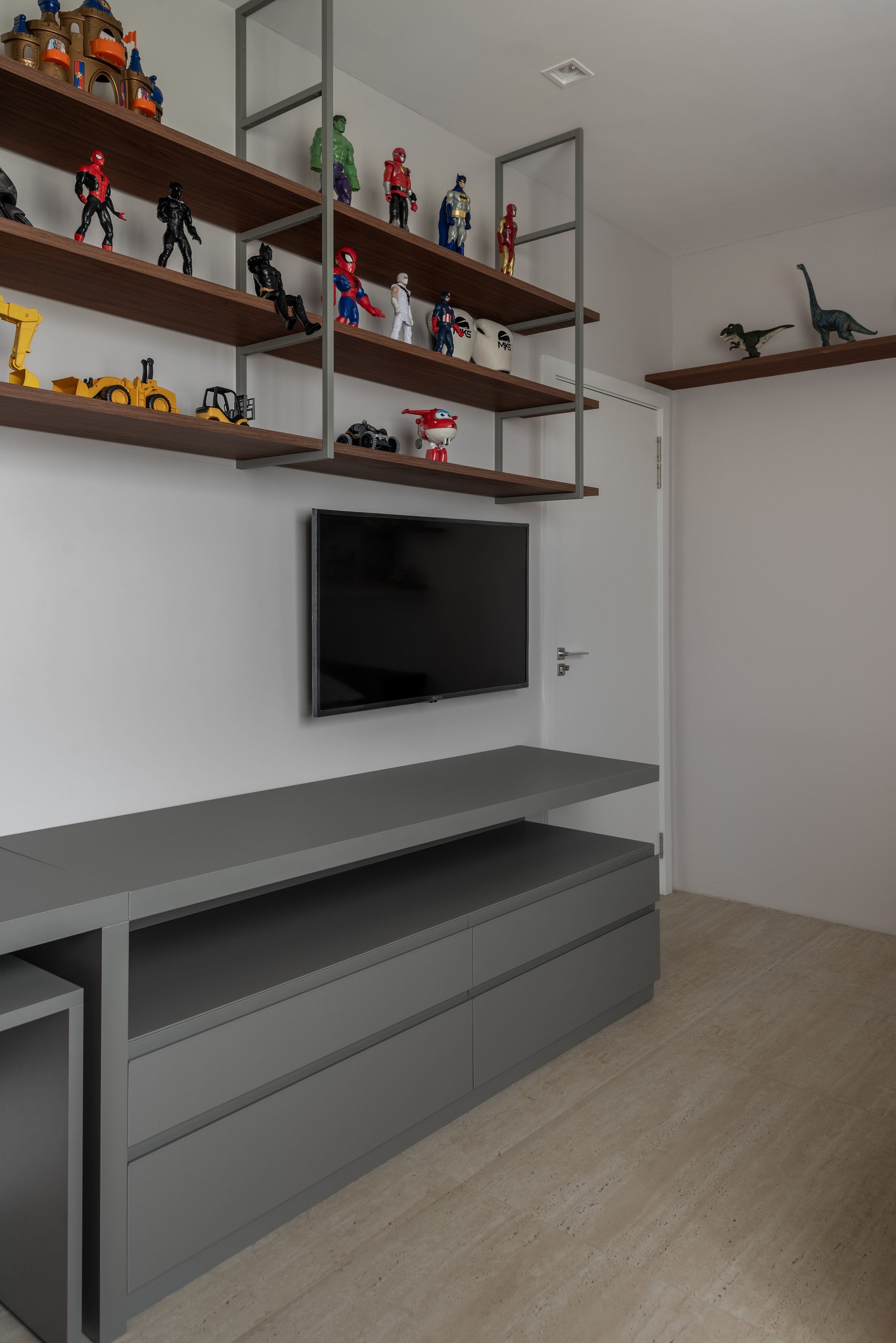 Wall-mounted shelf with action figures and toy models of superheroes and construction vehicles, television below, and a gray cabinet.