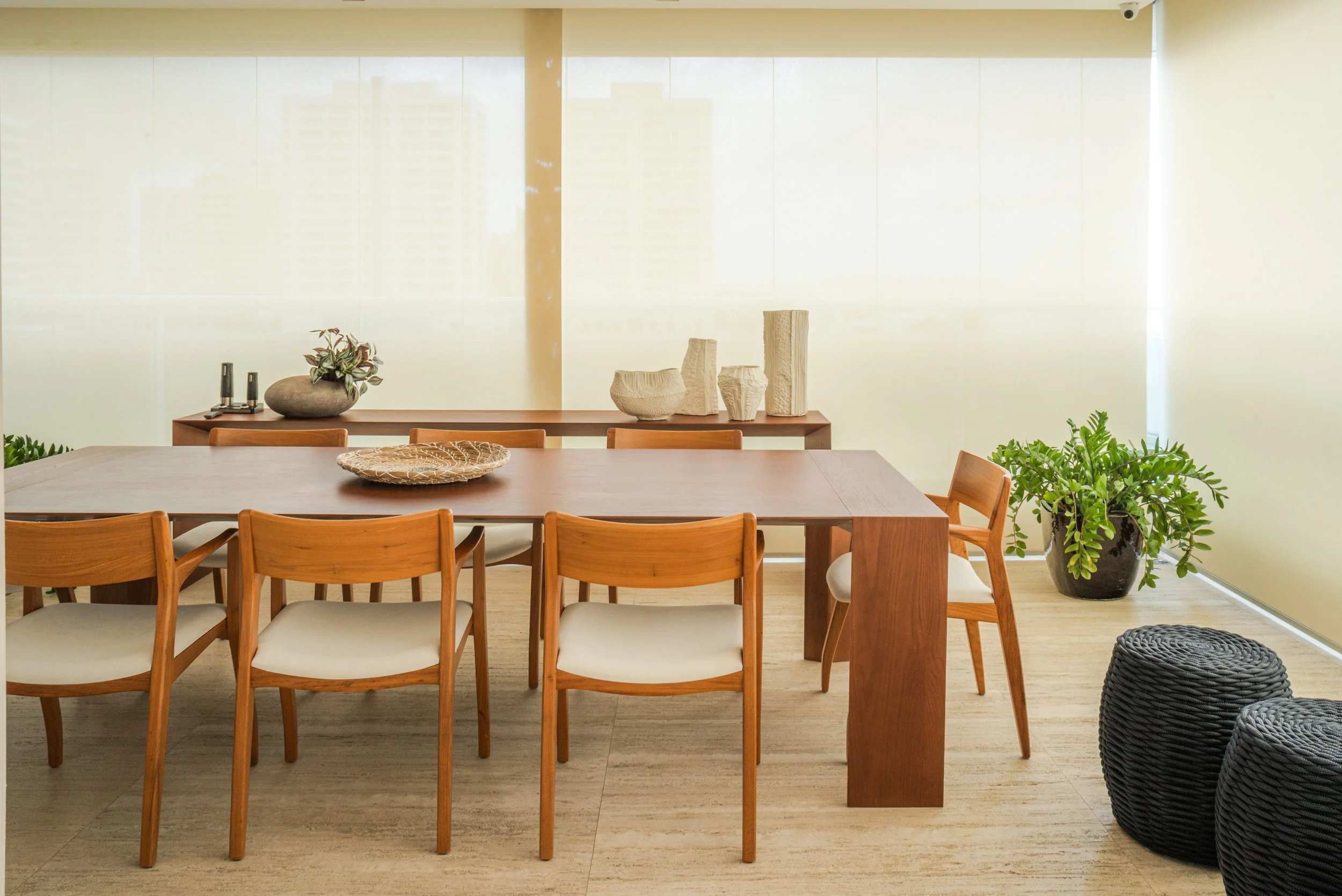 Minimalist dining room with a large wooden table surrounded by eight wooden chairs with white cushions. On the table, there is a decorative woven tray. In the background, a sideboard holds a potted plant, black decorative objects, and carved white sc