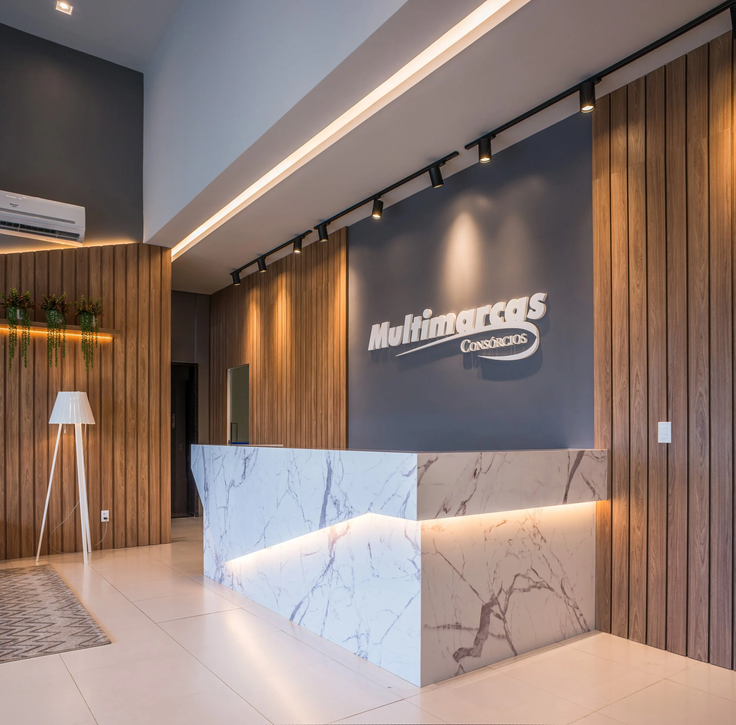 Modern office reception area with wooden wall panels, a marble reception desk, and the sign 'Multimarcas Consórcios' on the wall. There is a tall floor lamp, a small rug, and some plants on a shelf.